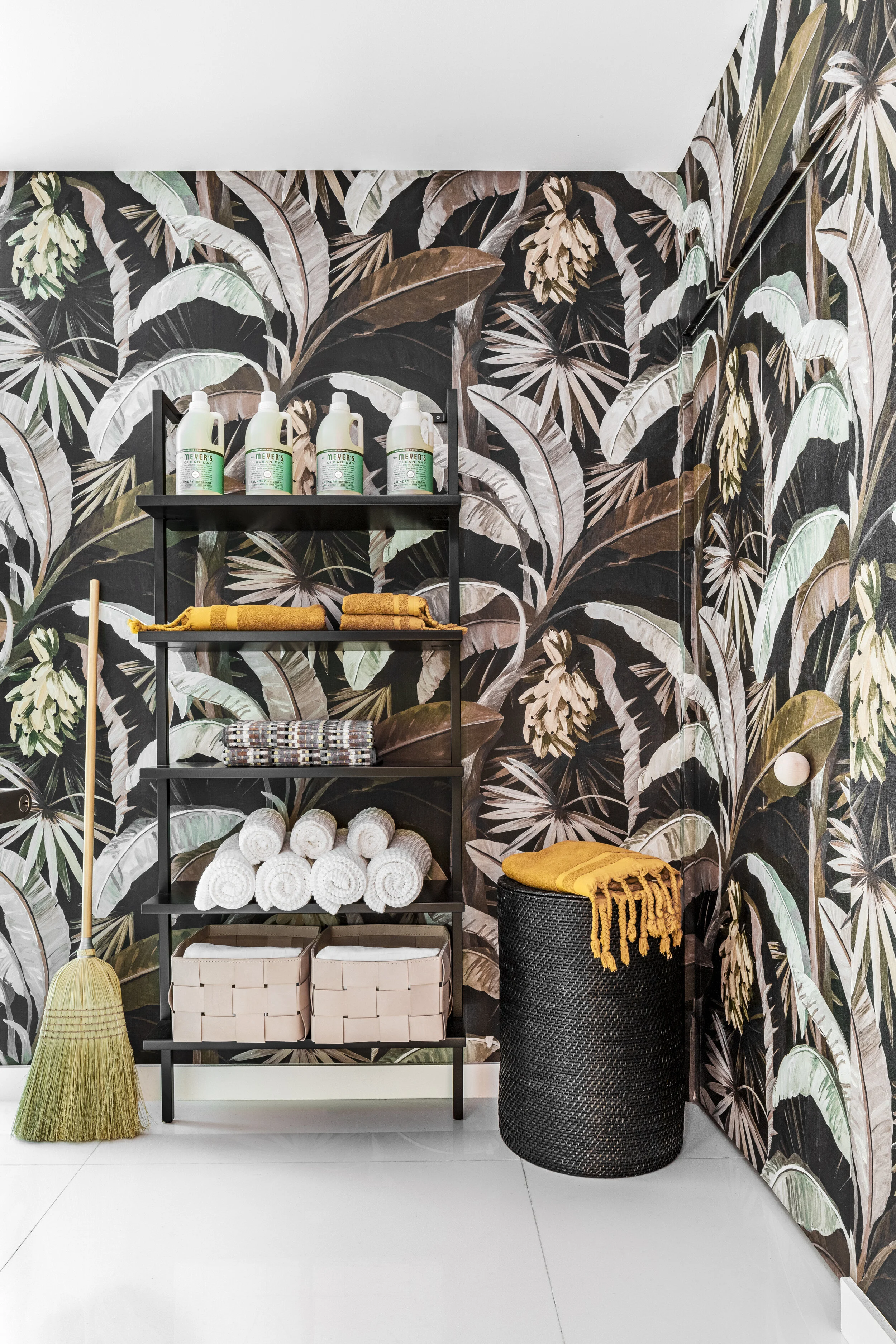 Shelving unit with cleaning supplies, towels, and baskets in a room with tropical leaf-patterned wallpaper. A broom leans against the wall, and a laundry basket with a yellow blanket is nearby.