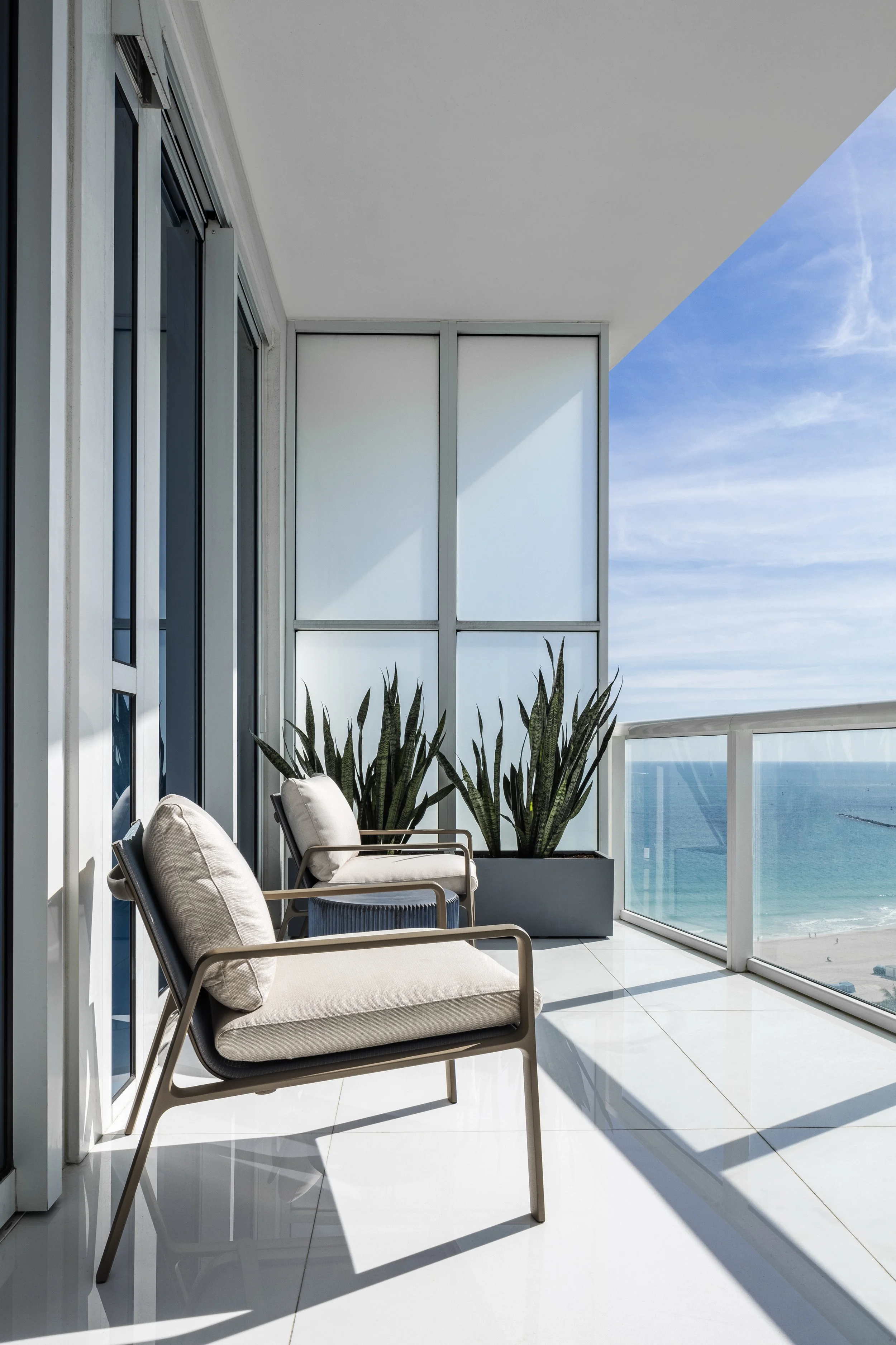 A balcony with two modern beige chairs, a large potted plant with green leaves, frosted glass panels, and an ocean view under a clear blue sky.