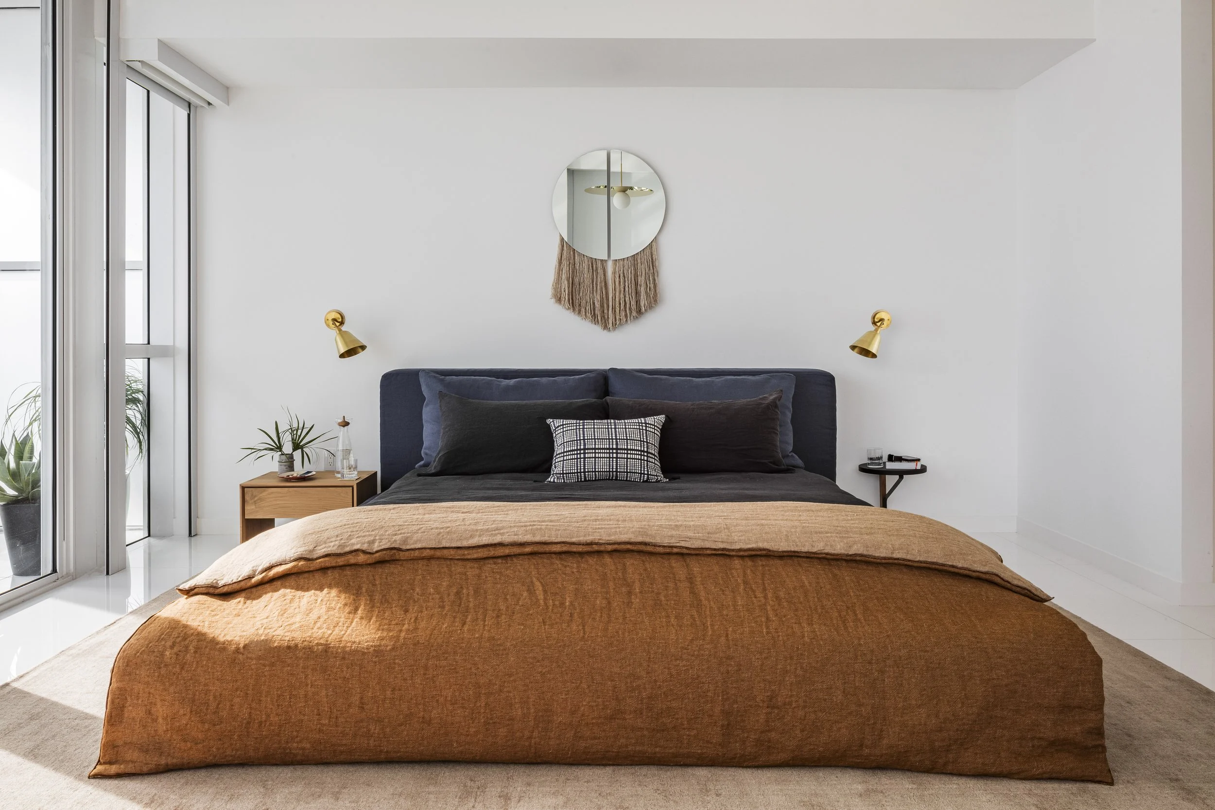 Modern bedroom with a large bed, dark pillows, a textured brown blanket, wall-mounted reading lights, a mirror with a woven wall hanging above, a wooden nightstand with plants, and large glass doors leading to a balcony.