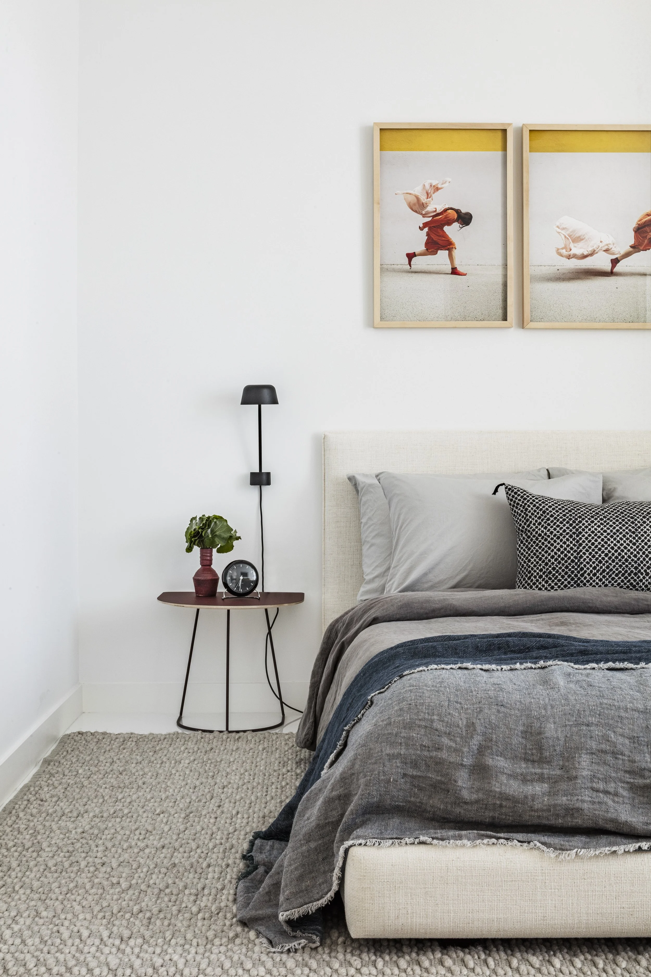 A bedroom with a beige bed, gray bedding, and black and white pillows. A small wooden nightstand holds a potted plant, a black round clock, and a black wall-mounted lamp. Two framed pictures of a girl running, with one image showing her mid-stride an