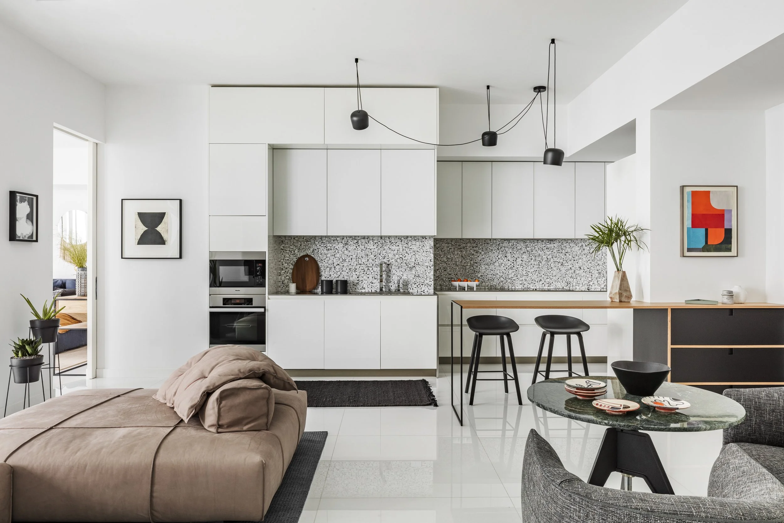 Modern open-plan living space with a kitchen featuring white cabinets, a black and white mosaic backsplash, black pendant lights, and a breakfast bar with two black stools. There is a beige sofa, a round green marble dining table with plates, and gra