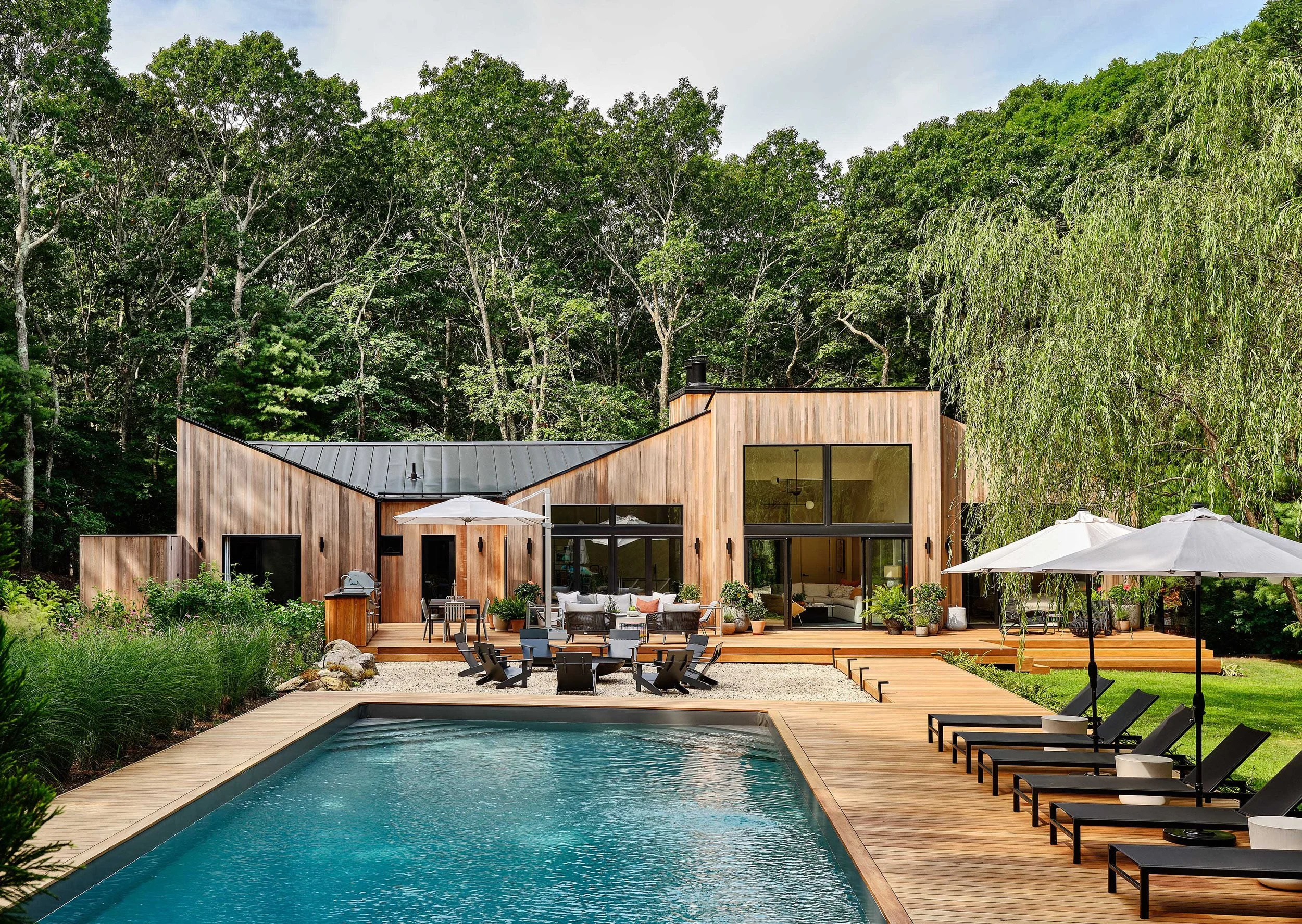 Modern house with wooden exterior, large glass windows, patio with outdoor furniture, swimming pool with lounge chairs and umbrellas, surrounded by lush green trees and greenery.