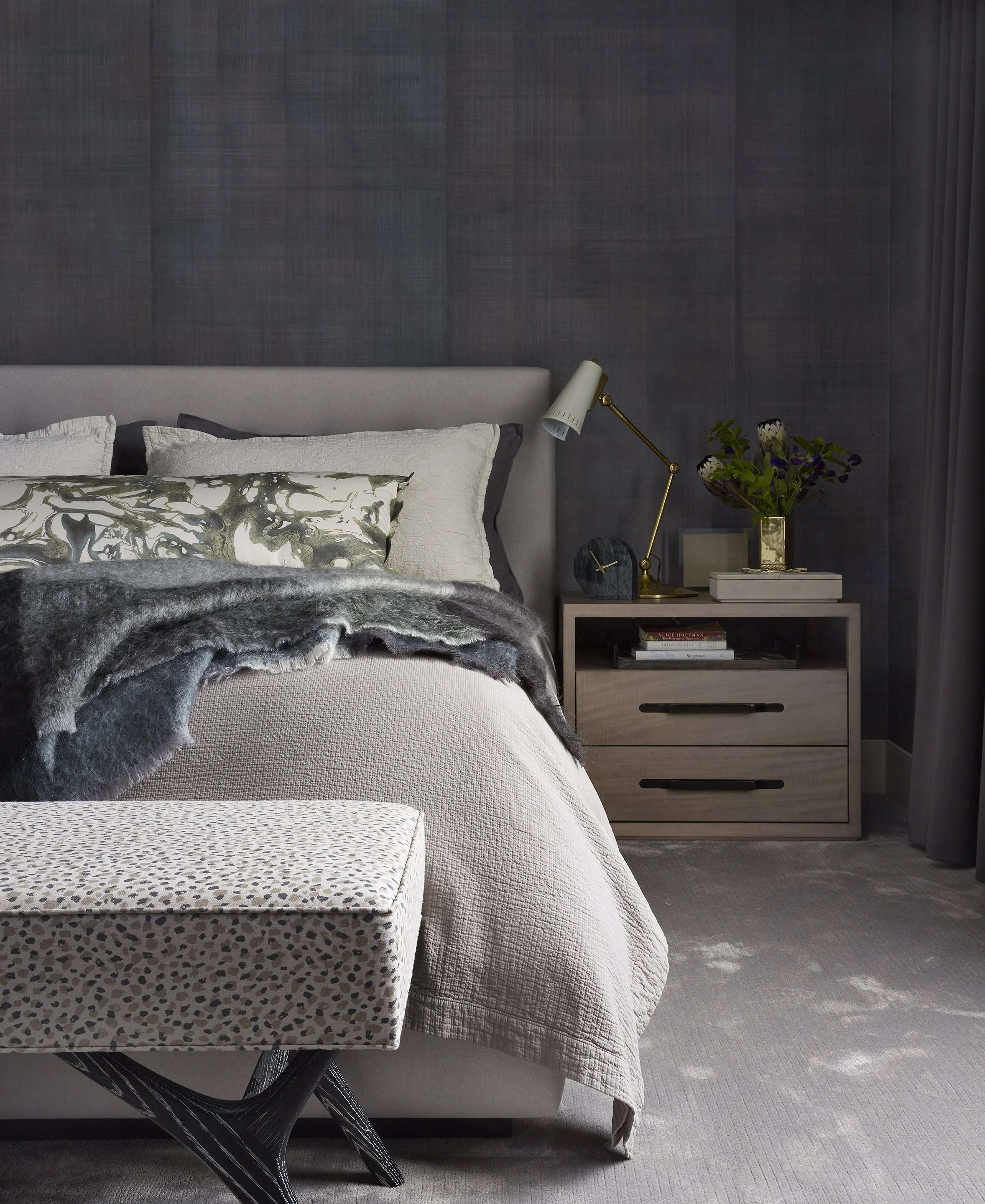 A bedroom with a bed, decorated with pillows and a throw blanket, a nightstand with a desk lamp, a clock, books, and a flower vase, with a textured gray wall and curtains.