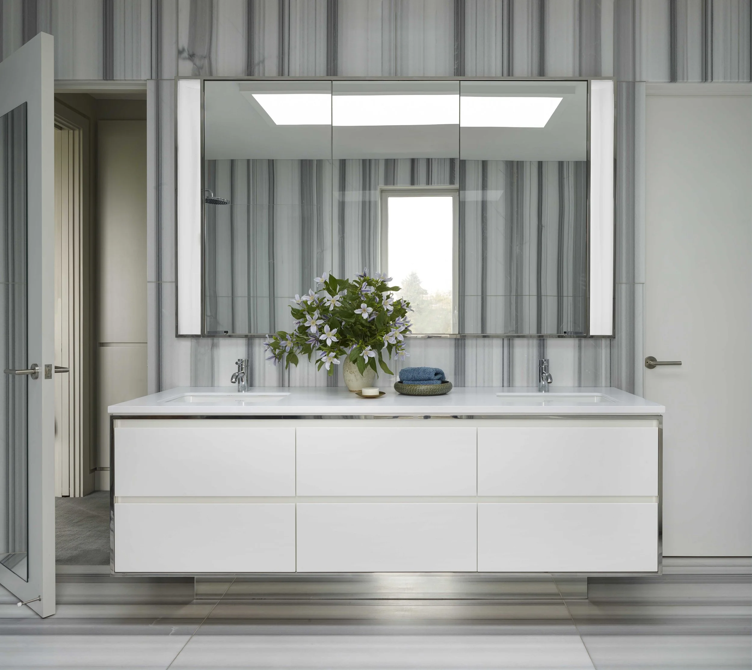 Modern bathroom with double vanity, large mirror, and gray marble tiles. Decorative vase with flowers and folded towels on the counter.