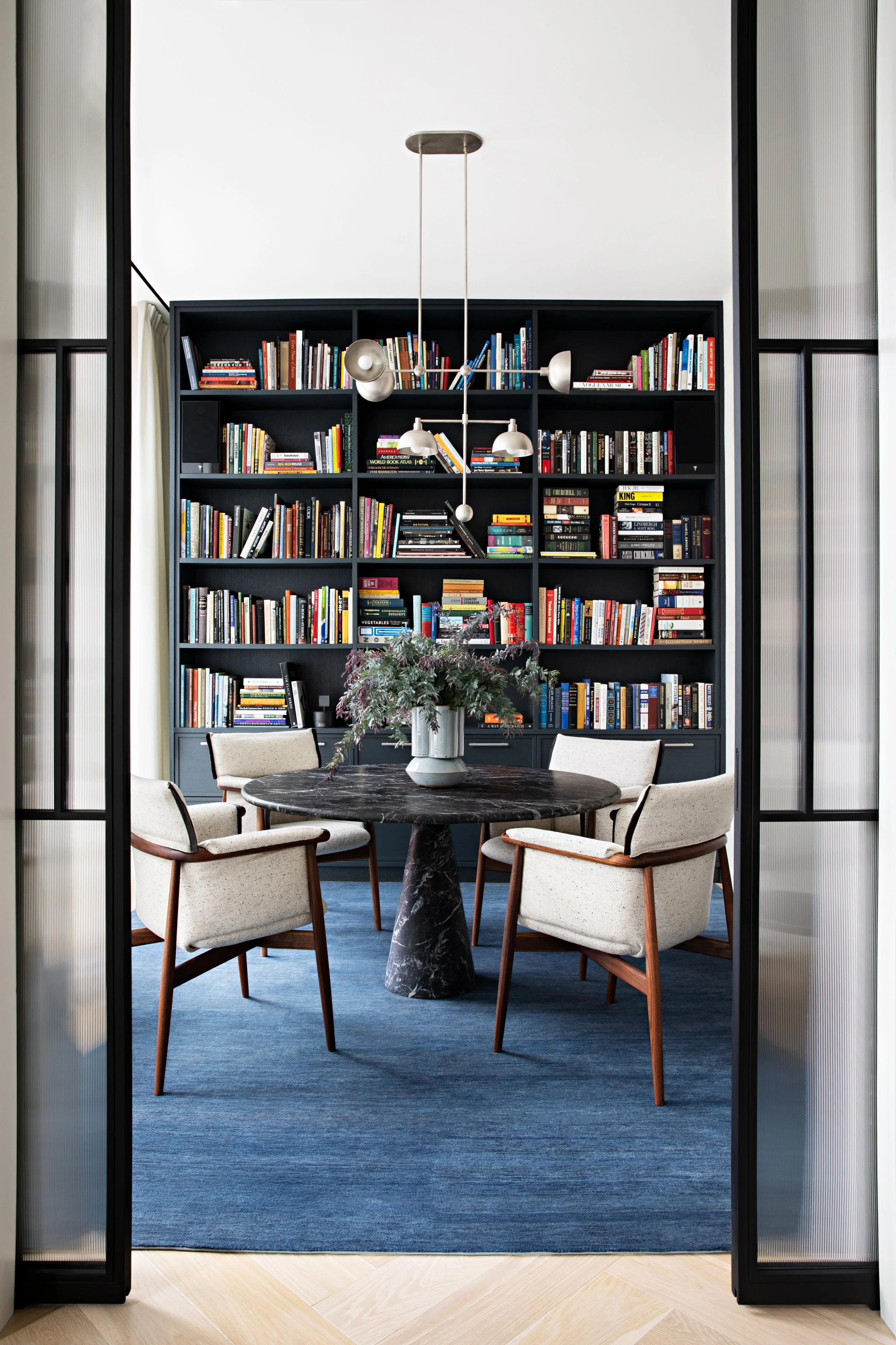 View through sliding doors into a dining area with a round black marble table and four beige upholstered chairs. A large blue rug covers the floor. A tall black bookshelf filled with books stands against the back wall. A modern chandelier hangs above the table, and a vase with flowers is on the table.