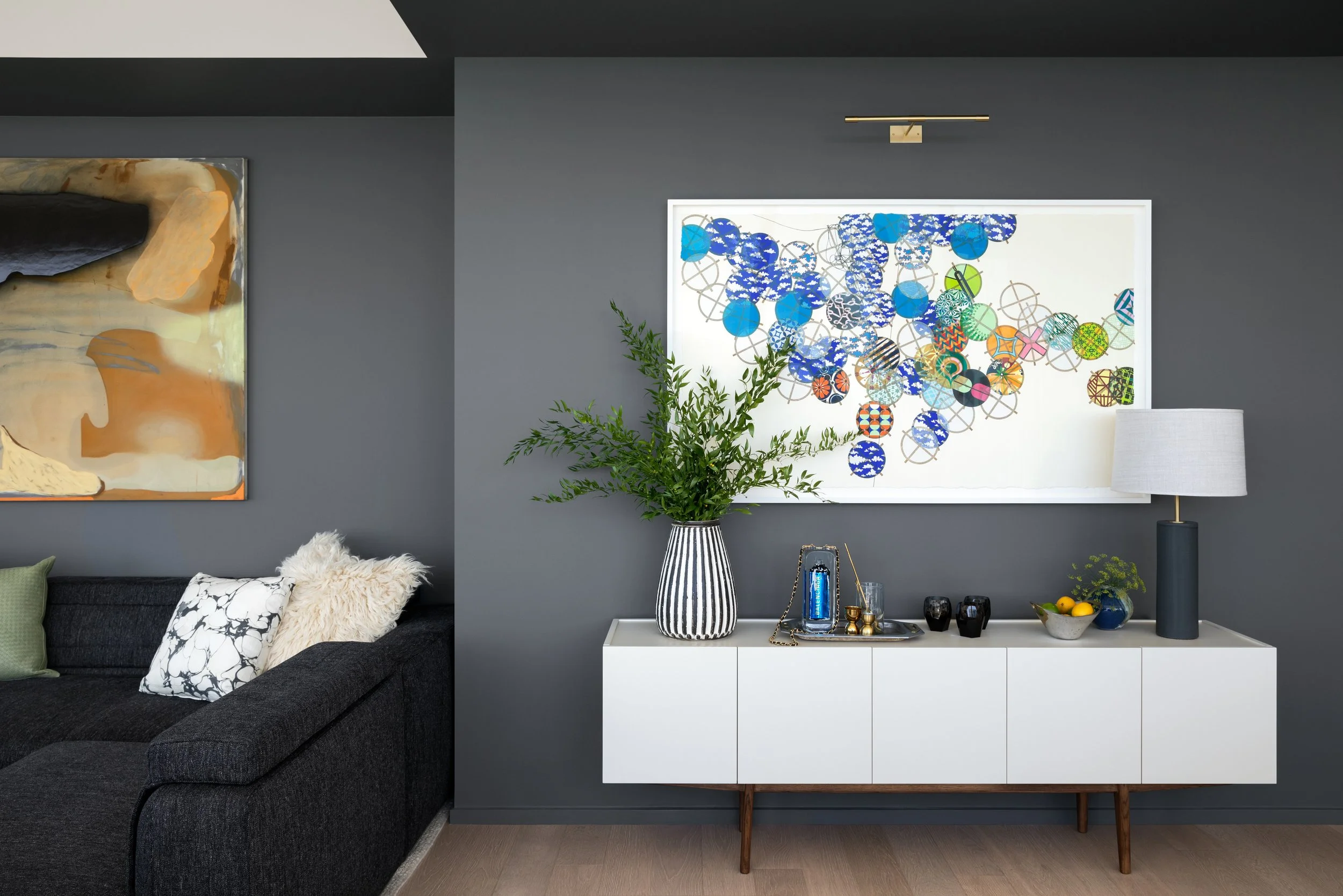 Interior of a living room with a dark gray wall, a white sideboard decorated with a large plant in a striped vase, a lamp, and decorative items, with abstract artwork hanging above the sideboard.
