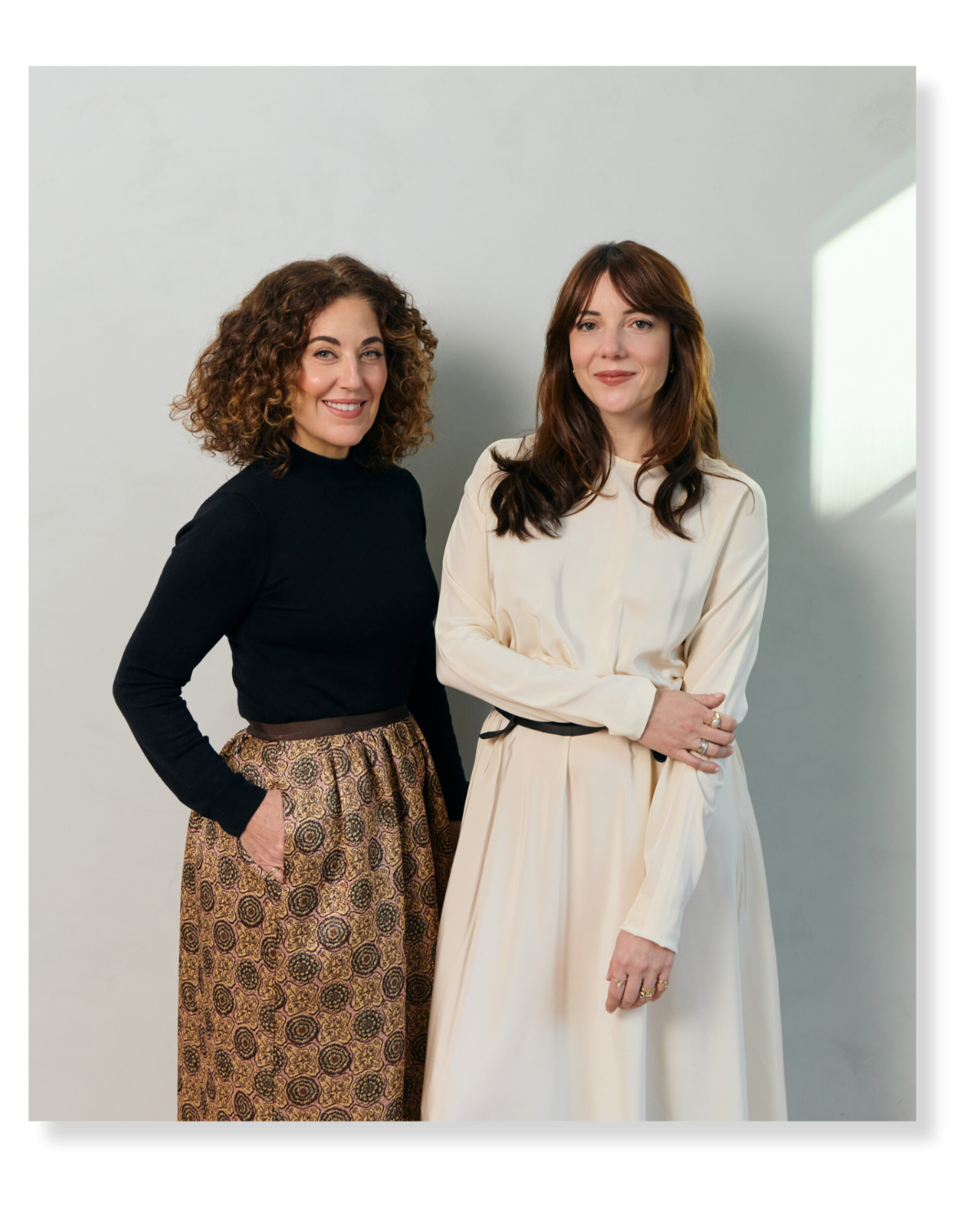 Two women standing against a plain wall, one with curly hair wearing a black top and patterned pants, the other with straight hair wearing a cream dress.