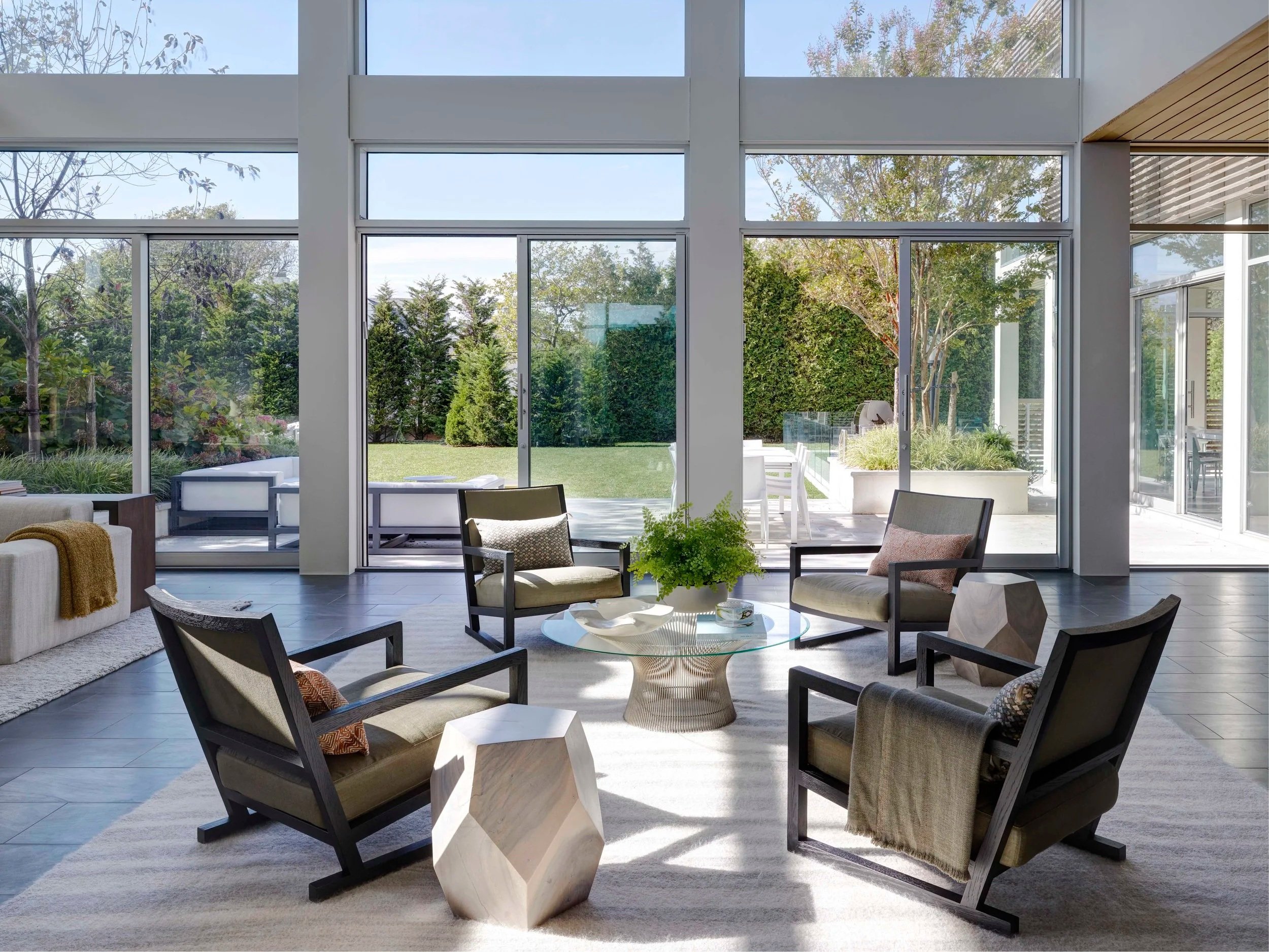 Modern living room with large windows, white walls, a sectional sofa with pillows, a glass coffee table, a mid-century armchair, and a balcony above.