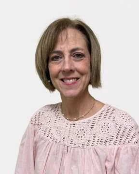 A woman with shoulder-length light brown hair smiling, wearing a light pink blouse with intricate crochet details on the upper chest and shoulders, against a plain white background.