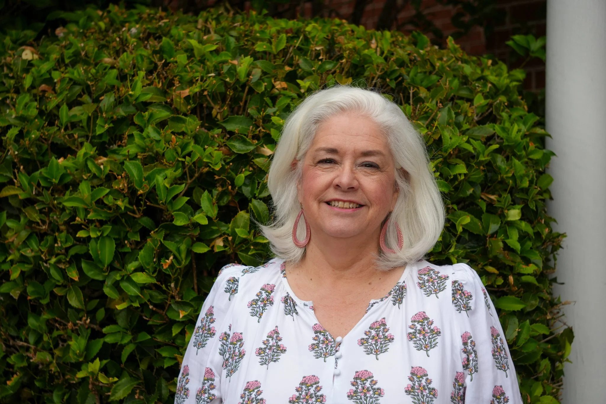 A portrait of a woman with shoulder-length gray hair, smiling, wearing a purple and pink patterned blouse against a plain white background.