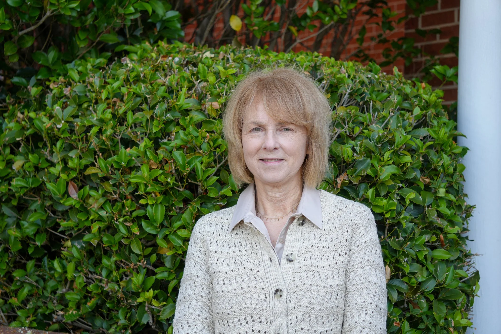 Portrait of an elderly woman with short blonde hair, wearing a light blue cable-knit sweater, a white collared shirt, and a gold necklace with a cross pendant, against a plain white background.
