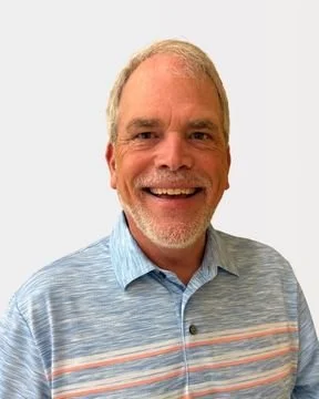 Smiling man with short gray hair wearing a blue collared shirt against a plain white background.