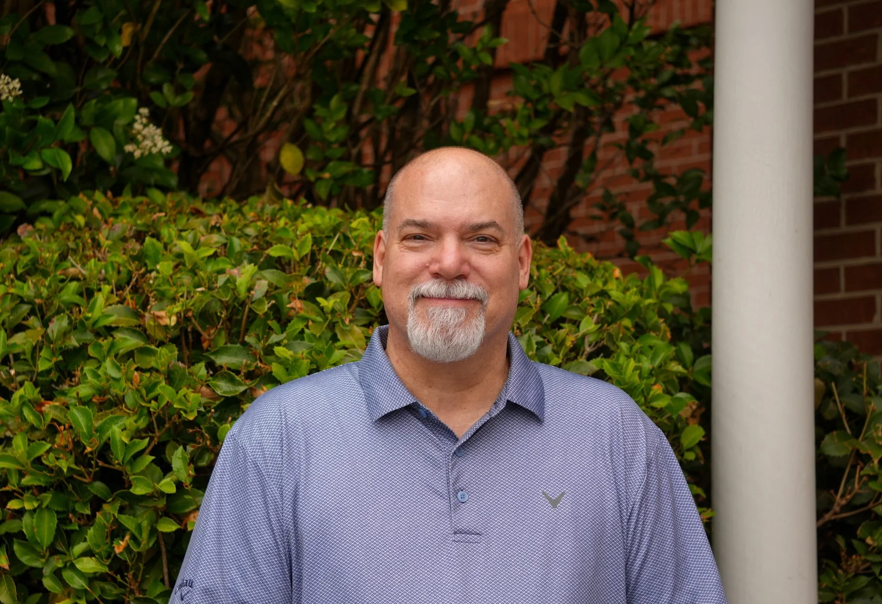 Headshot of a bald man with a gray beard, wearing a black collared shirt, smiling against a white background.