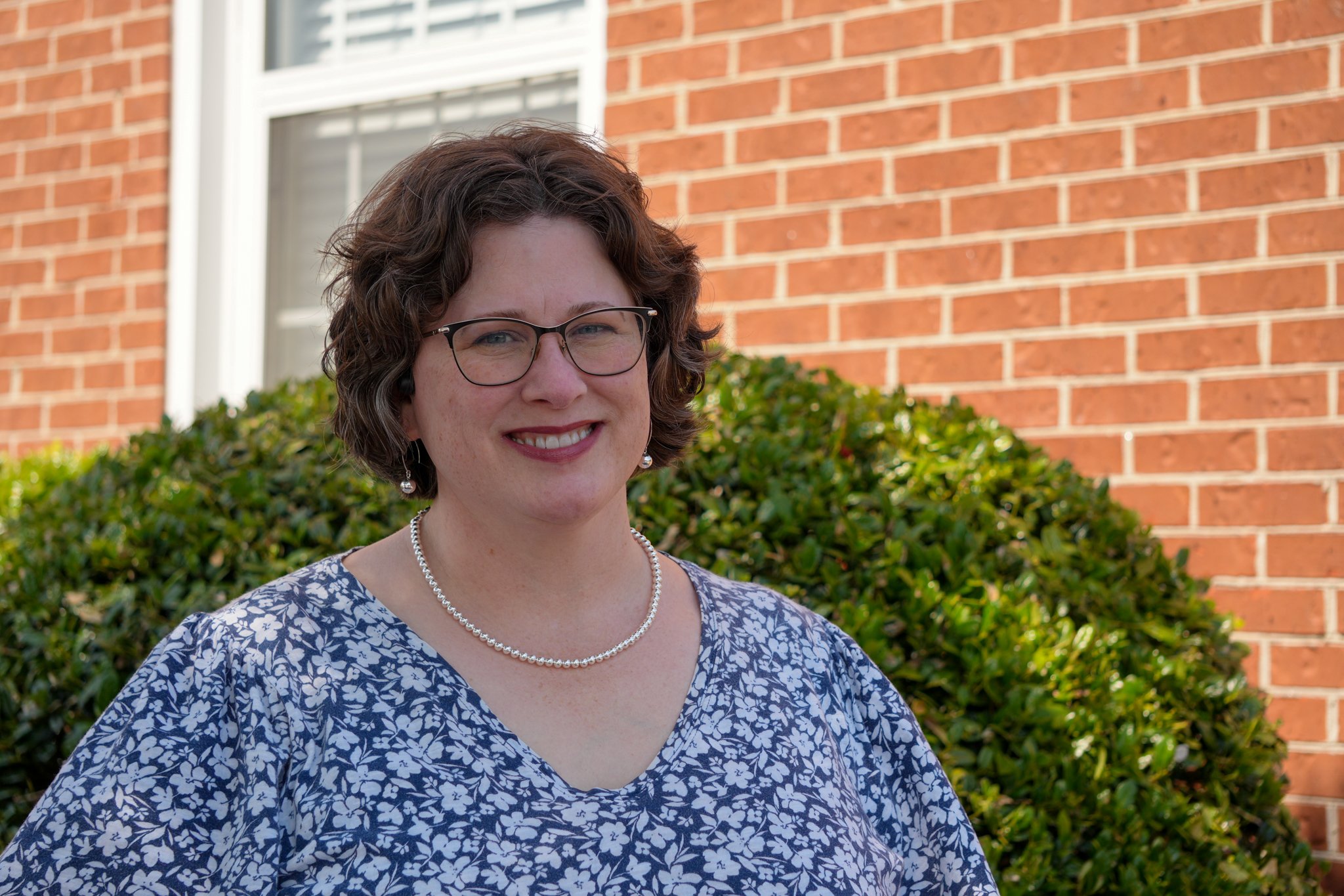 Smiling woman with short hair and glasses, wearing a blue floral blouse and pearl necklace, against a plain white background.
