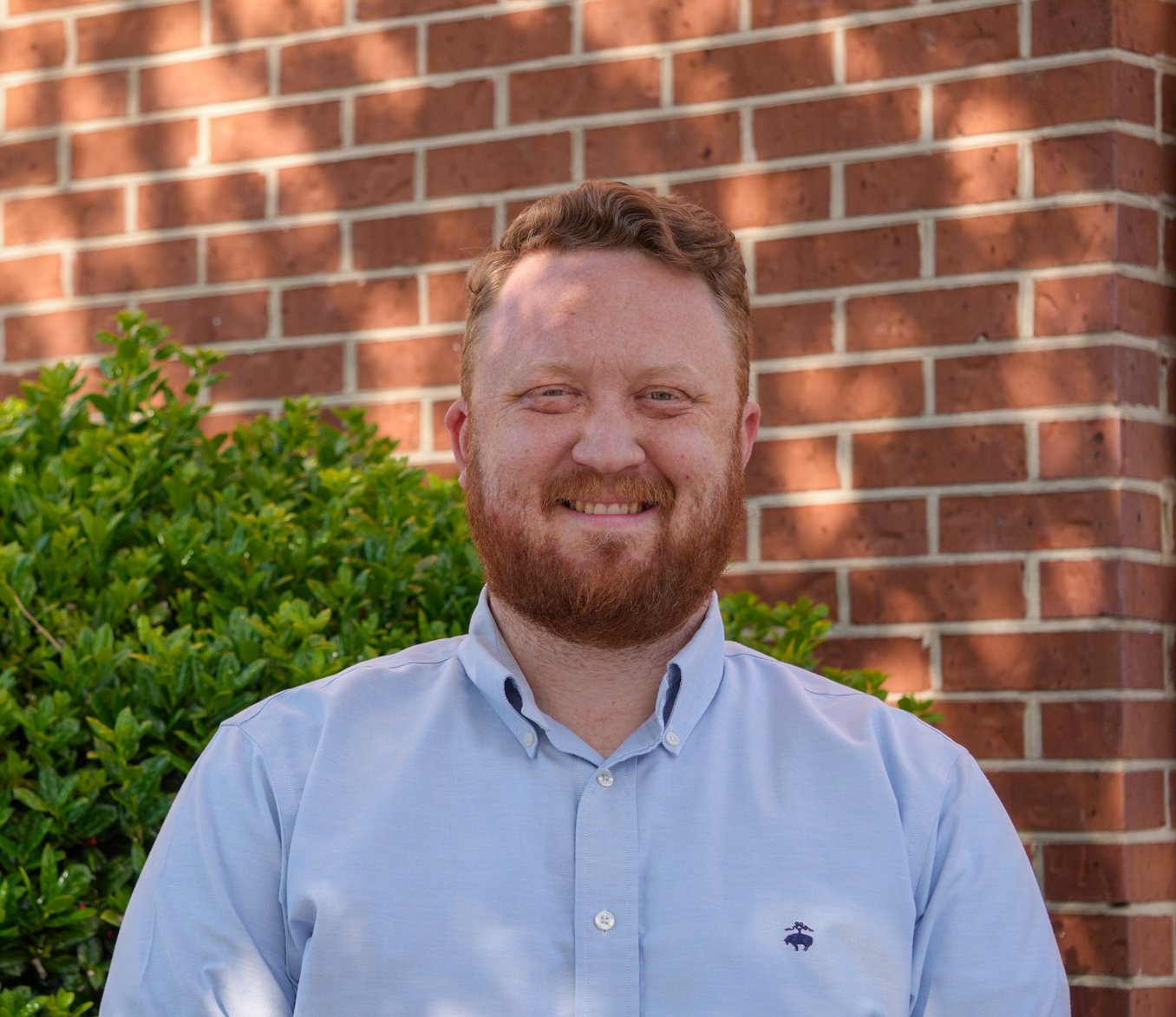 A man with a beard and short curly hair is wearing a light gray blazer and a blue dress shirt, smiling in front of a plain white background.
