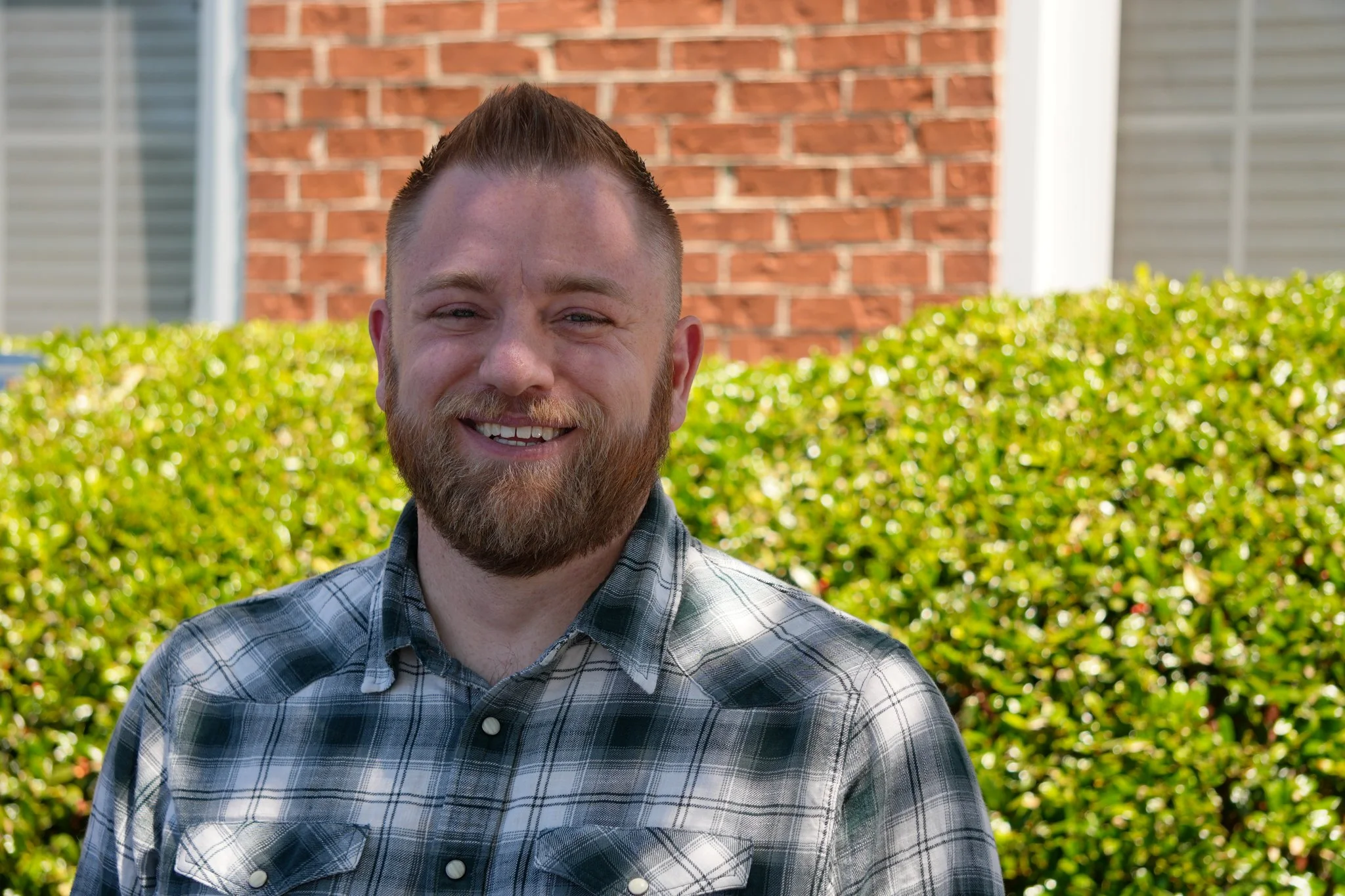A smiling man with a beard and short hair, wearing a gray button-up shirt, standing against a white background.