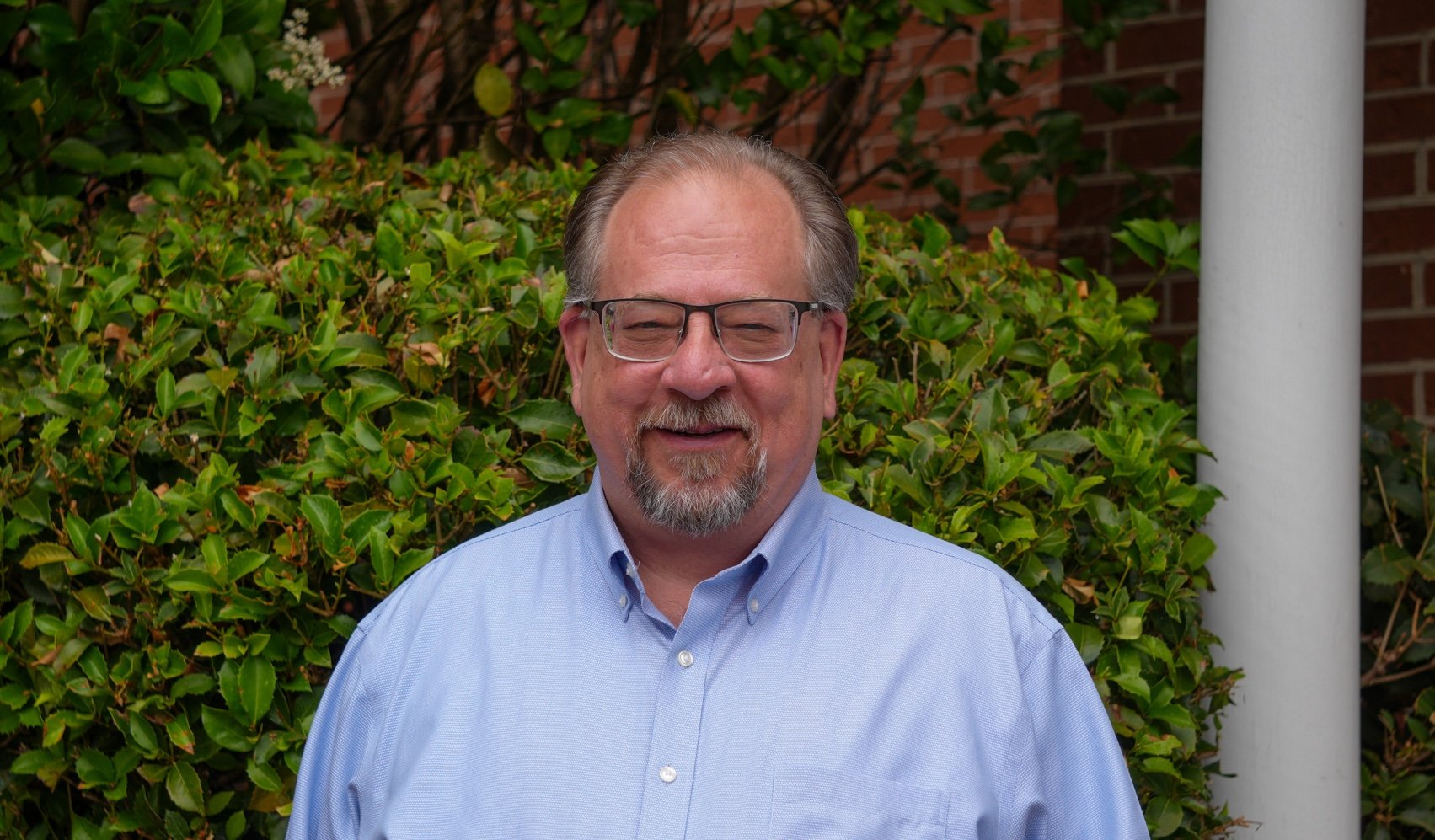 A smiling man with glasses and a beard, wearing a blue checkered suit jacket and white shirt, standing against a plain background.