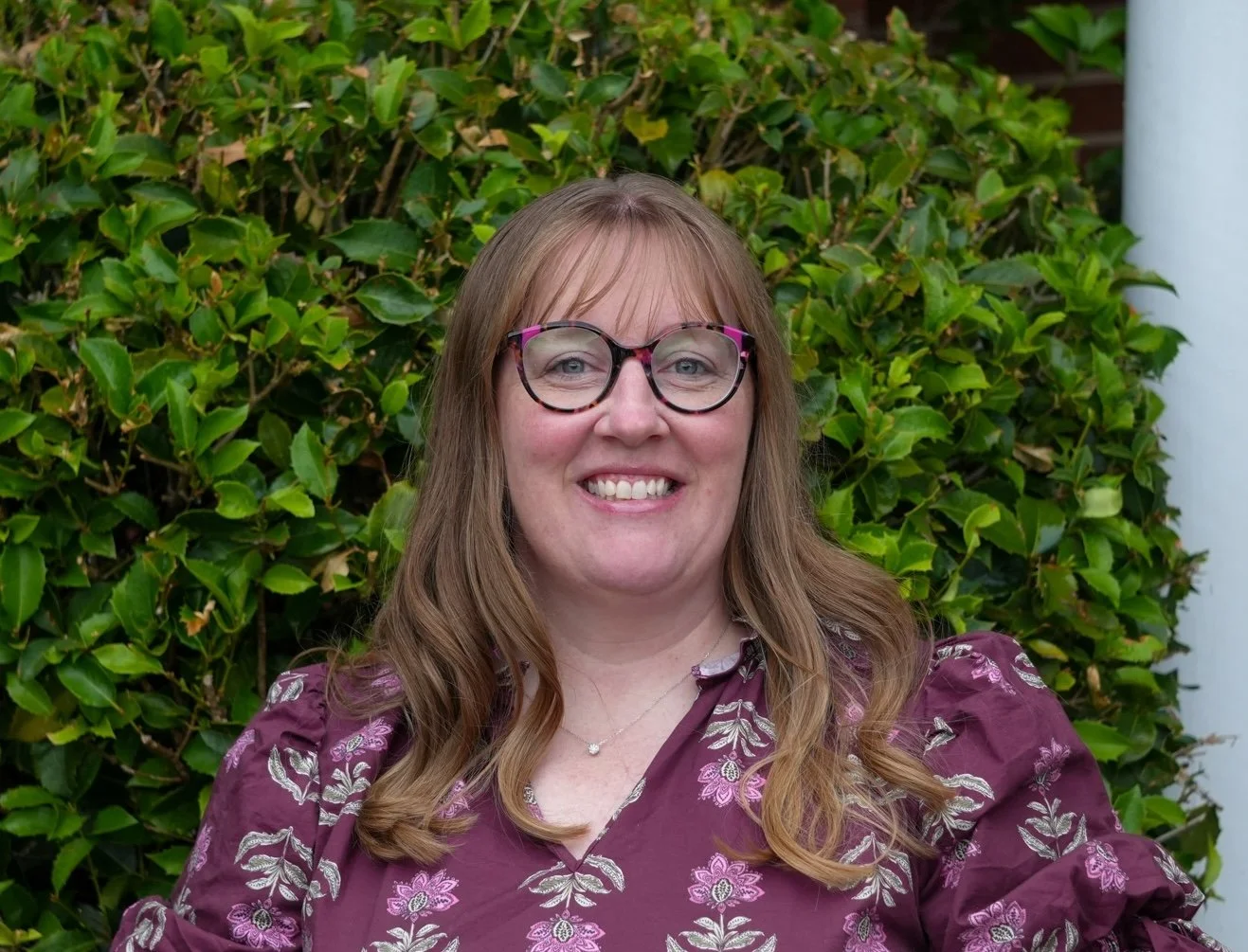 Headshot of a woman with shoulder-length light brown hair, wearing glasses and a floral top, smiling against a plain white background.