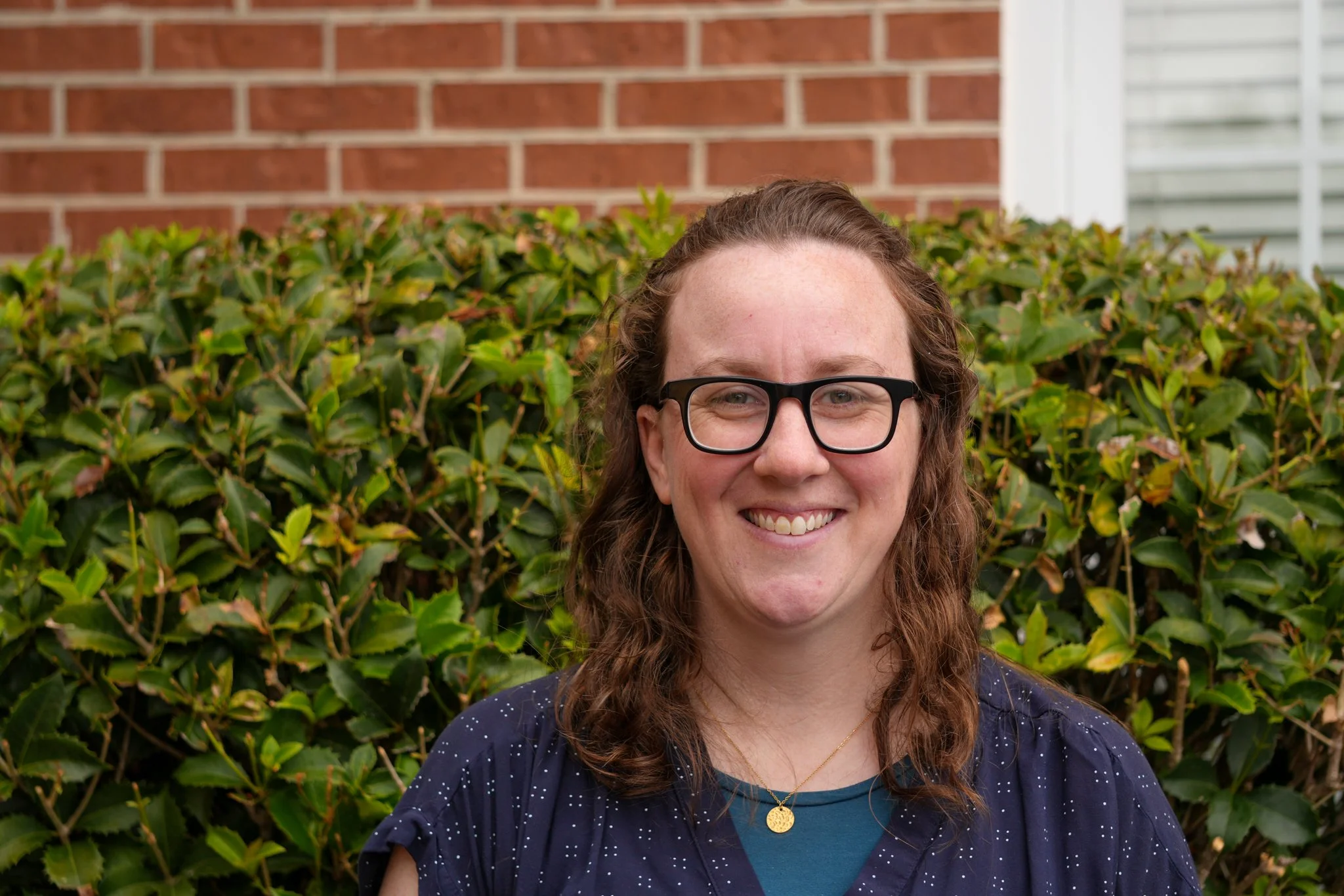 A woman with wavy brown hair, glasses, and a striped shirt smiling at the camera.