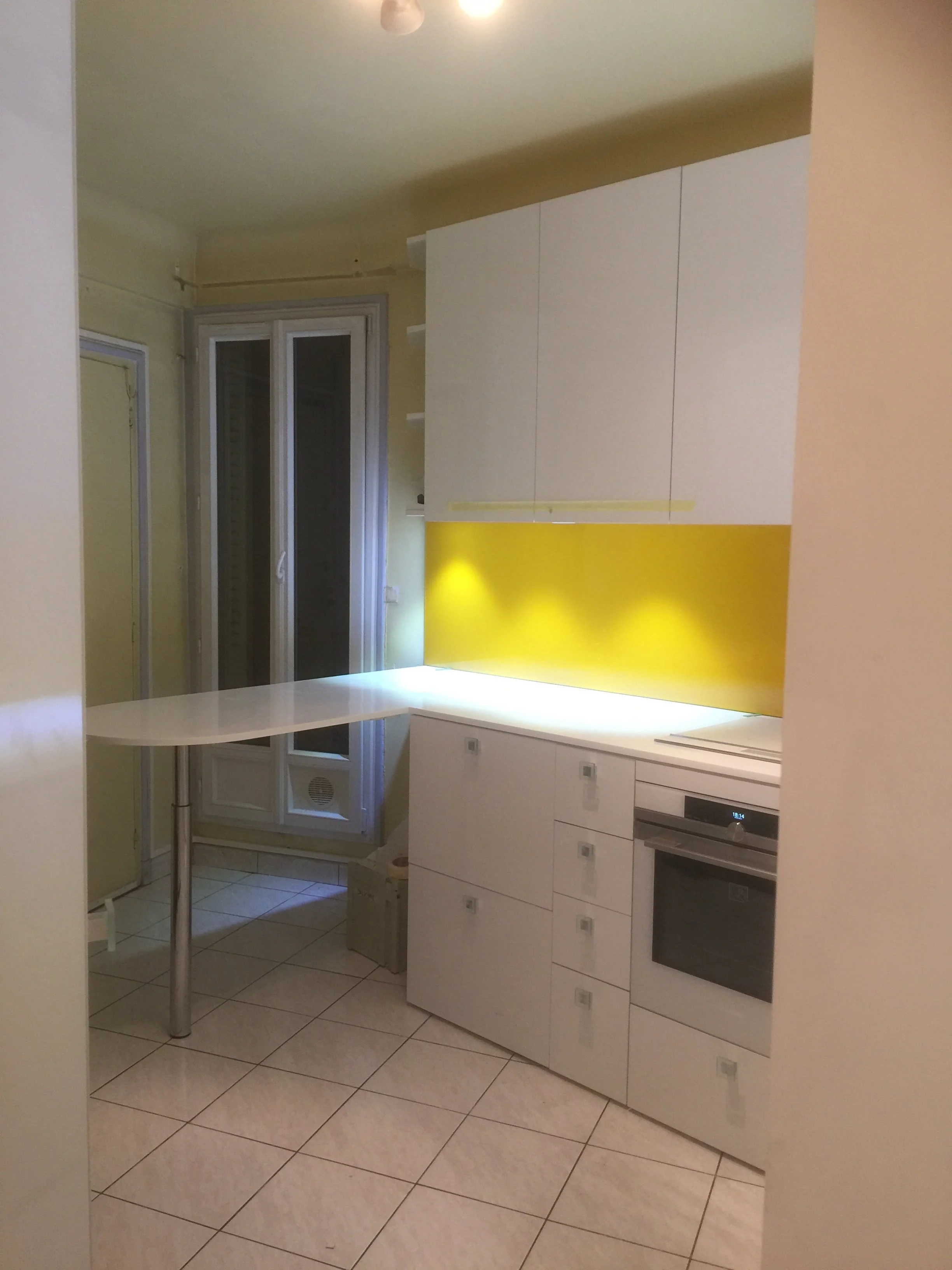 Modern kitchen with white cabinets, oven, tiled floor, and a yellow accent wall. A small table with a metallic leg is visible, and there is a large window with closed shutters.