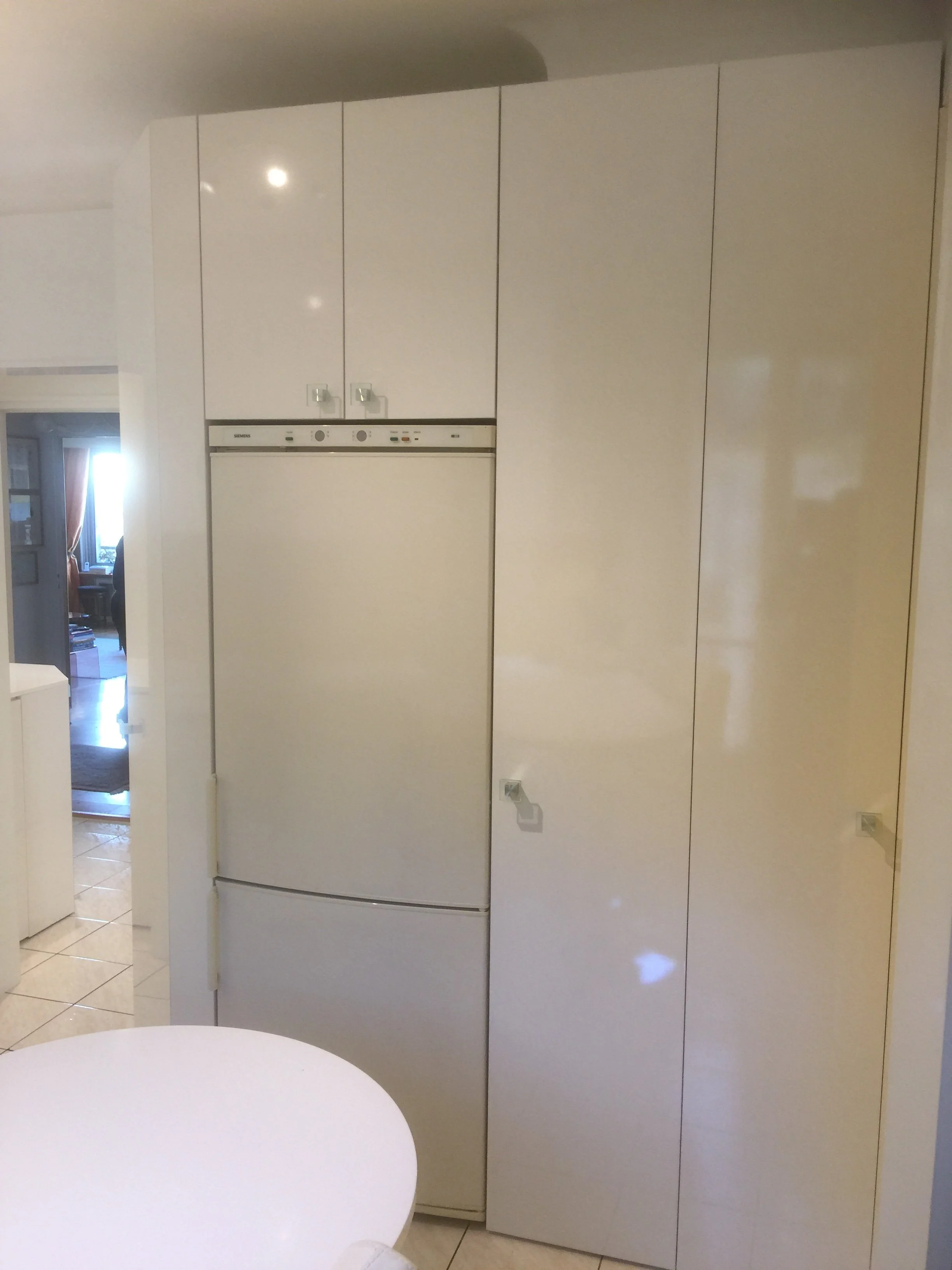 White kitchen cabinets with a small white refrigerator in the center, beige tiled floor, and a partial view of a round white table in the foreground.