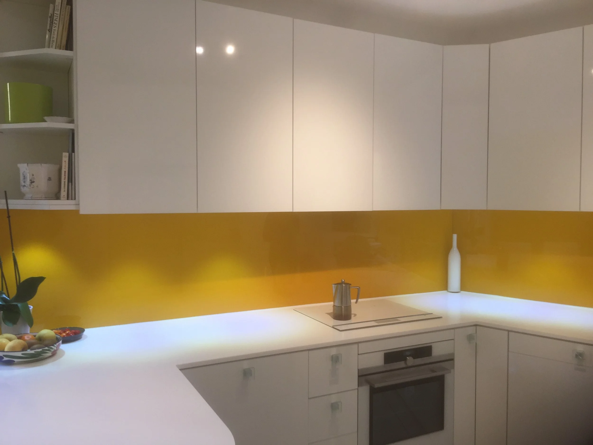 Modern kitchen with white cabinets, a yellow accent wall, a white countertop, a bowl of fruit, a tea kettle, and a white vase.