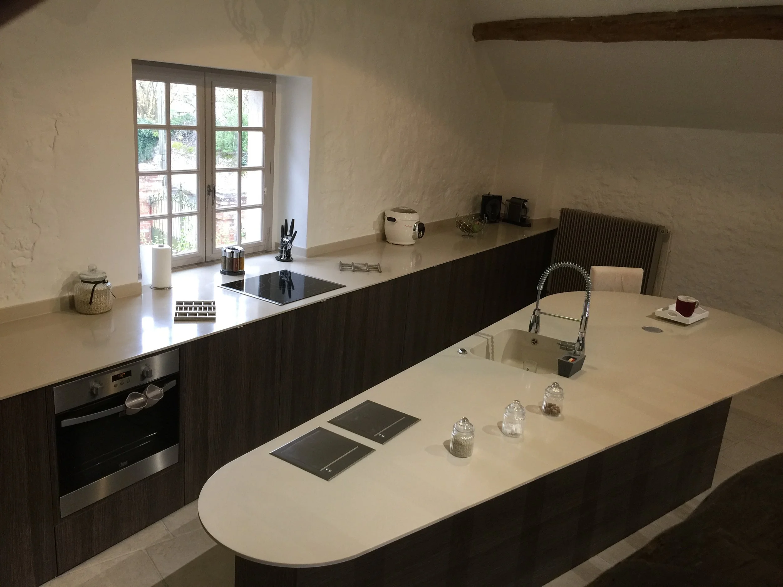 A modern kitchen with white countertops, dark cabinetry, and a gas stove. A window brings in natural light, revealing greenery outside. Items on the counter include a rice cooker, a fruit bowl, and kitchen utensils.