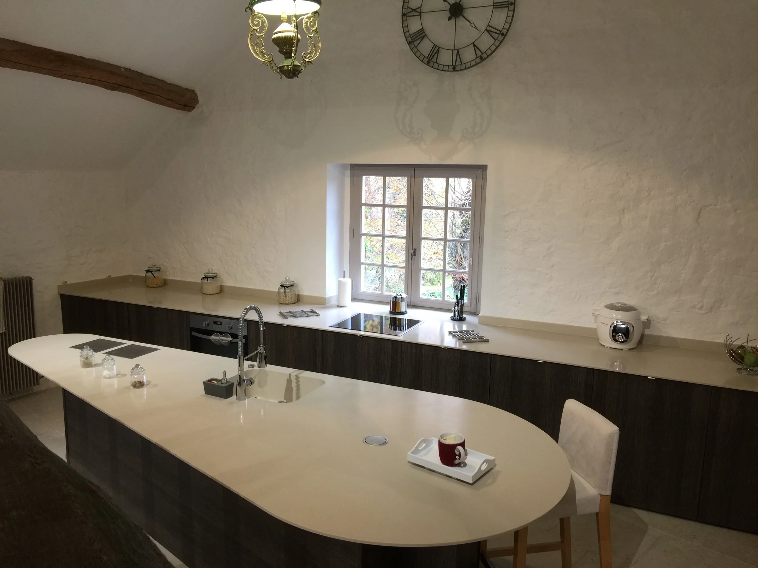 A kitchen with a large curved island, a window, and a clock on the wall. The island has various kitchen items including a soap dispenser, a mug, and a tray. The wall is painted white with a textured finish, and there is a wooden beam on the ceiling.