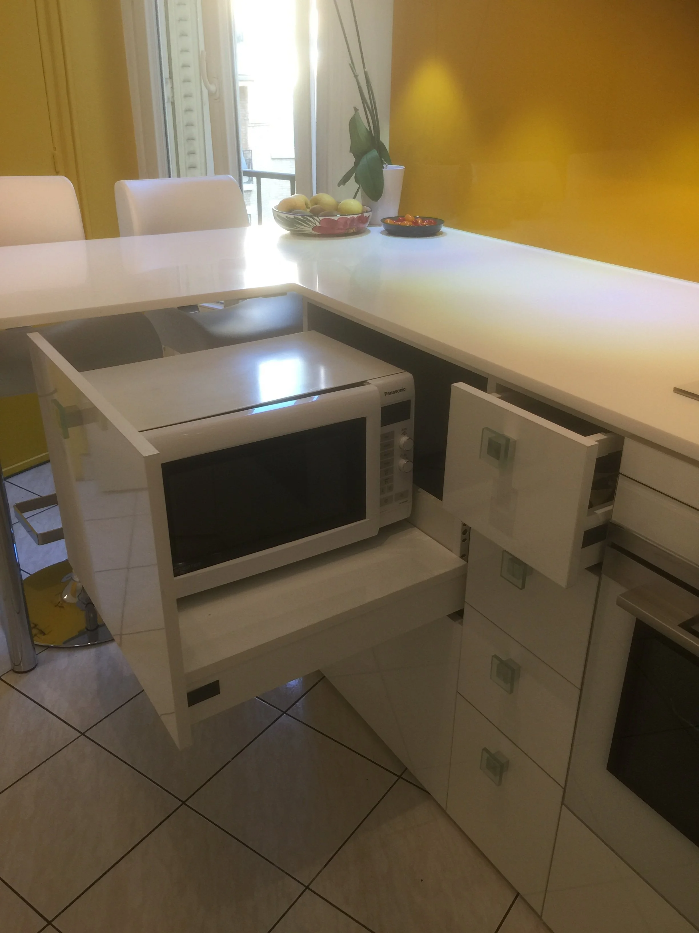 A dining room corner with white cabinets, a white table, and white chairs. On the table, there is a bowl of apples, a small dish of cherry tomatoes, and a potted plant near the window. The room has a yellow wall and tiled floor.