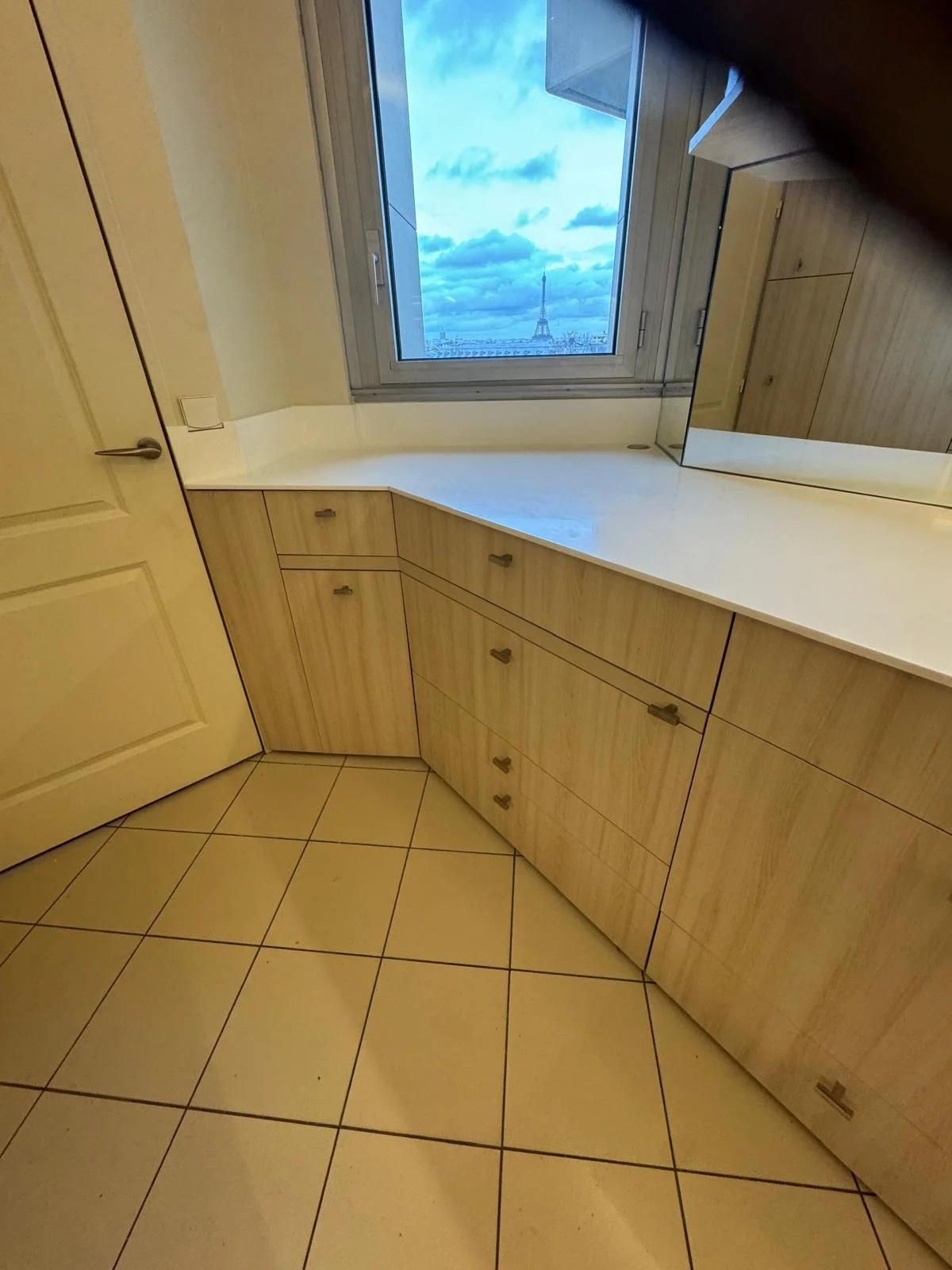 Indoor room corner with beige wooden cabinets, tiled floor, and a window with a view of the Eiffel Tower and cloudy sky.