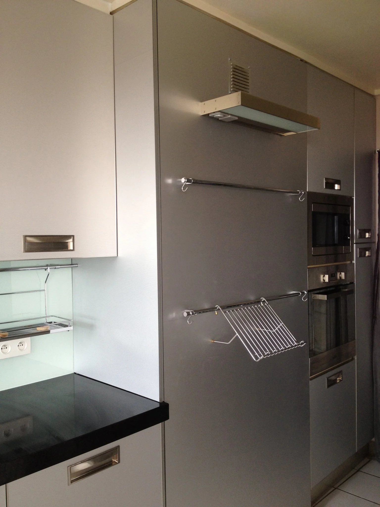 A kitchen wall with cabinets, a black countertop, a microwave oven, and a silver dish rack hanging from hooks on a wall.