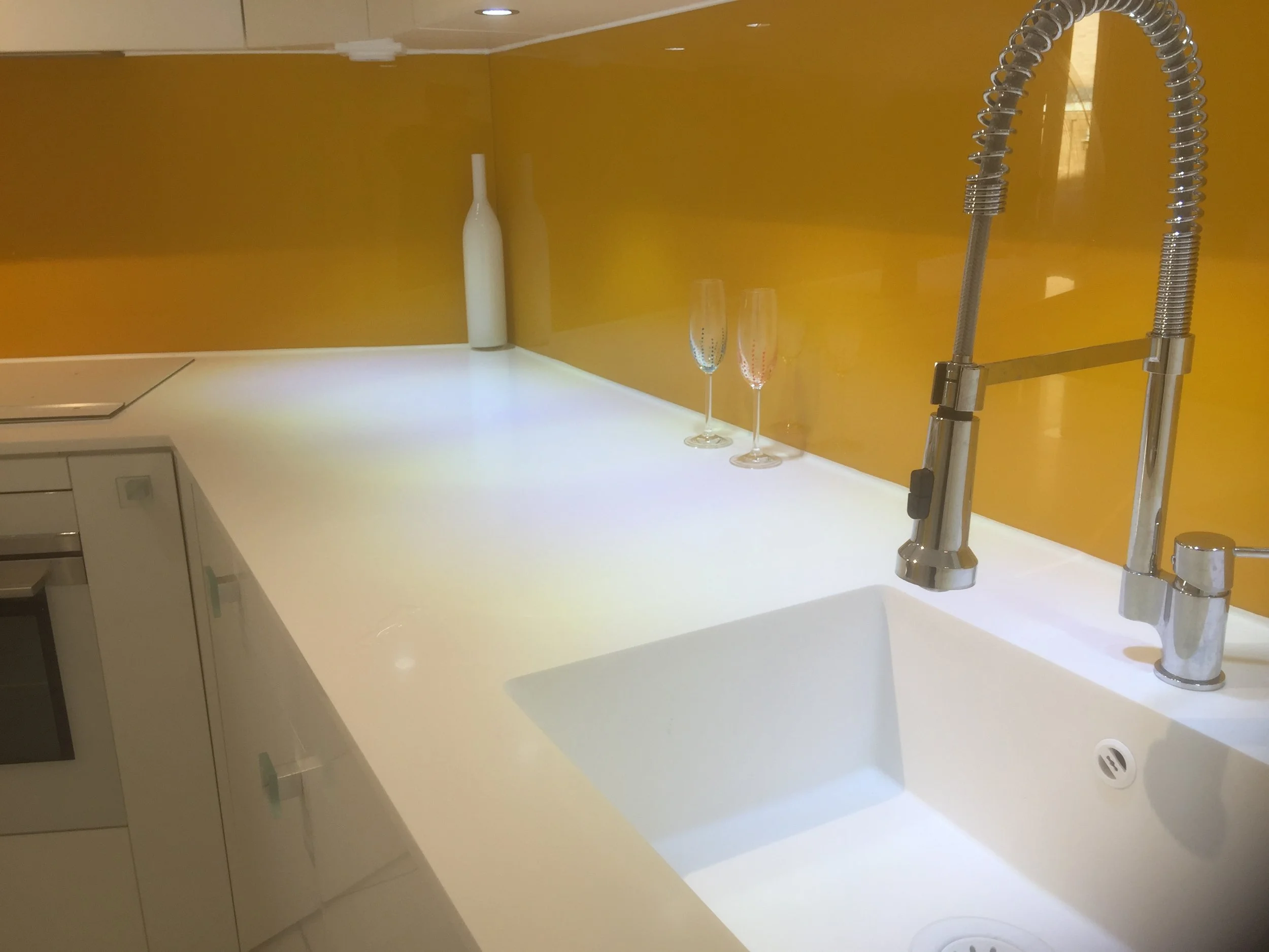 A modern kitchen counter with a yellow backsplash, a tall white vase, two champagne glasses, a metallic faucet with a spring spout, and a white drawer and cabinet.