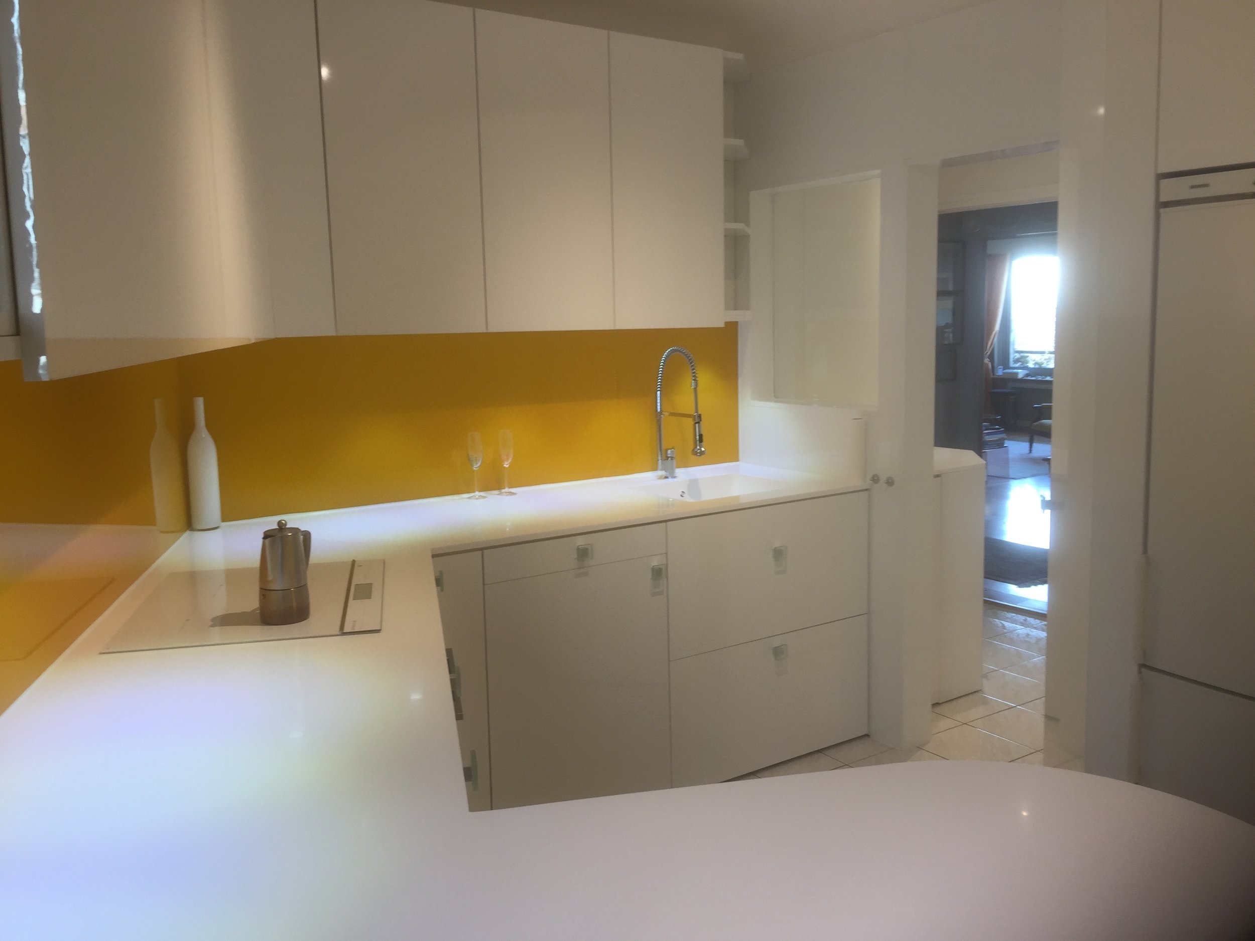 Modern kitchen with white cabinets, yellow backsplash, white countertops, and stainless steel faucet. Doorway leads to a room with a window and curtain.