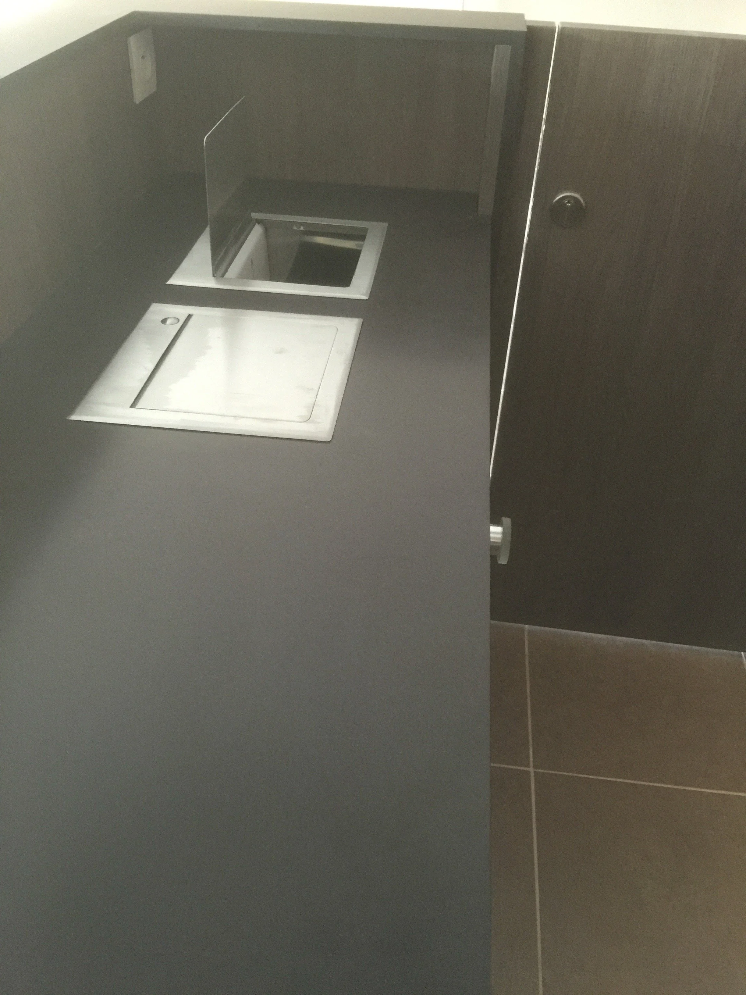 A dark gray bathroom stall with a white tilt-down seat cover and a small, open trash can embedded in the wall.