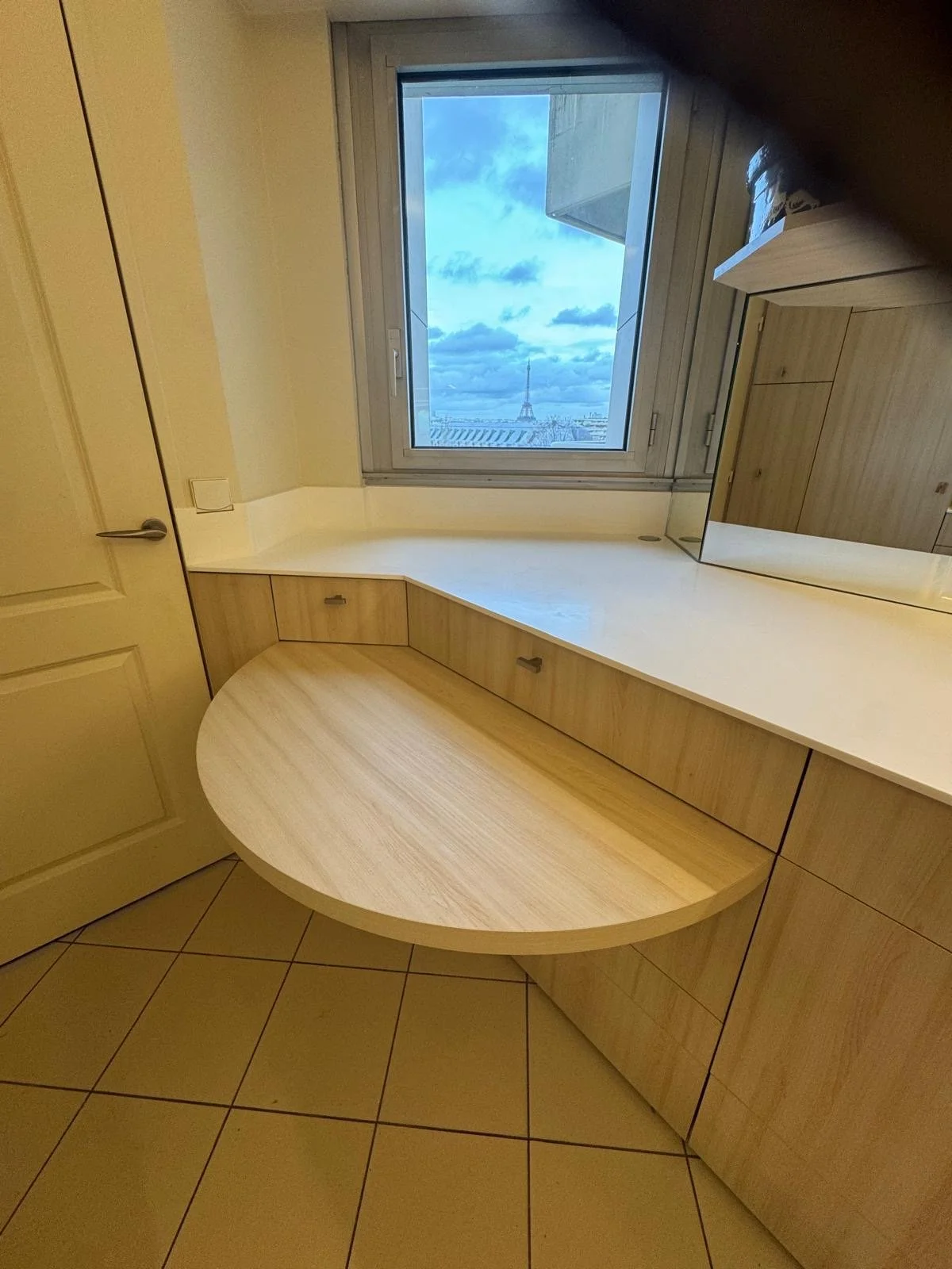 Room with wooden built-in desk under a window showing a Paris Eiffel Tower view, with beige tile floor and closed beige door.