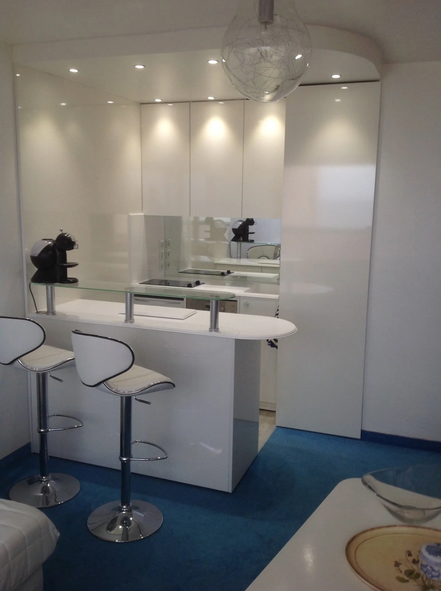 A modern kitchen with white cabinets, a small countertop, black kitchen appliances, and three white barstools with chrome bases around a high white table. The room has white walls and a blue carpet. There is a mirror reflecting the kitchen area, a sp