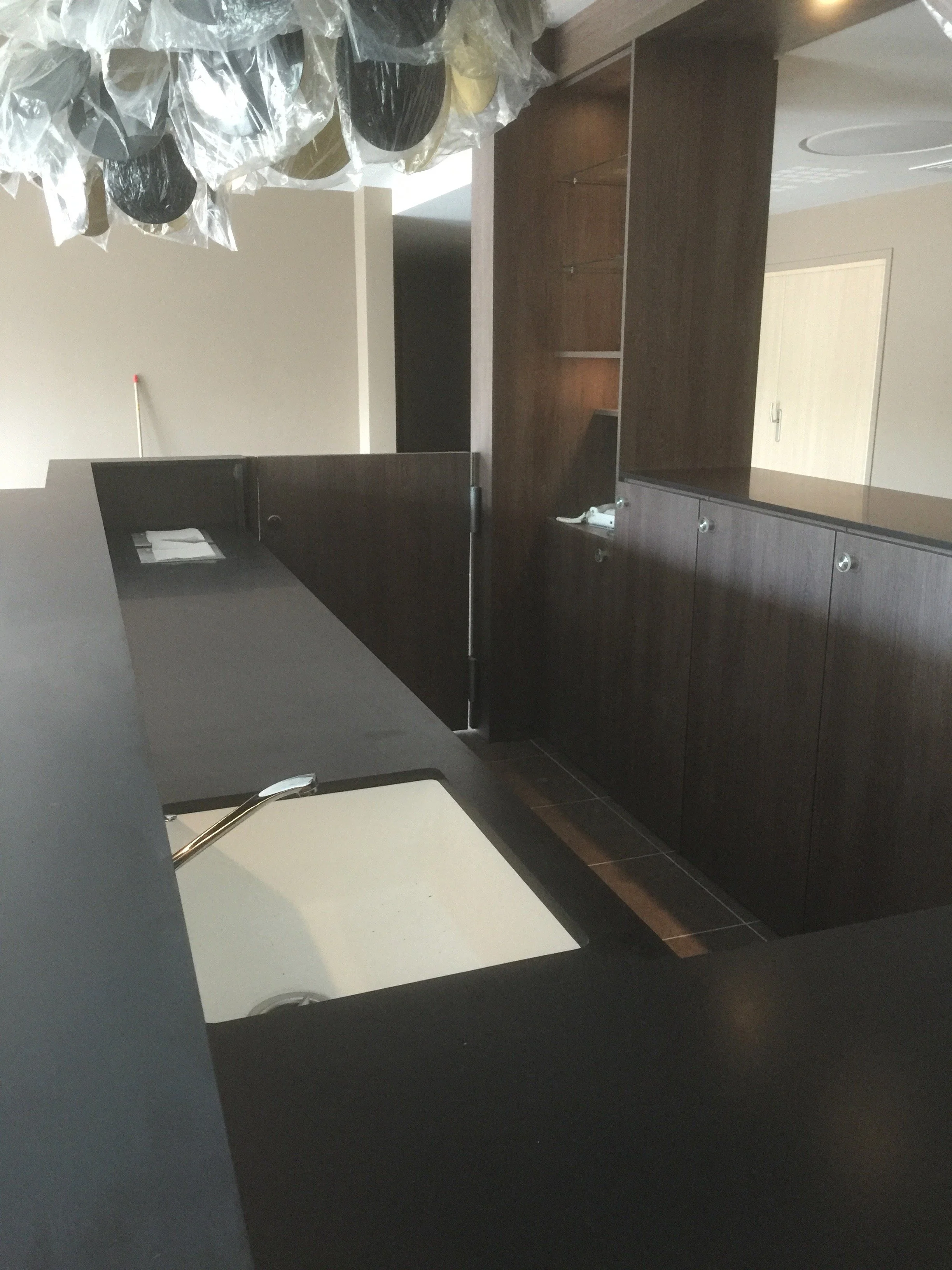 A kitchen countertop with a dark surface, a white cutting board, a metallic faucet, a dark wooden cabinet with open shelves, a window with a handle, and a section of a wall with a light switch.