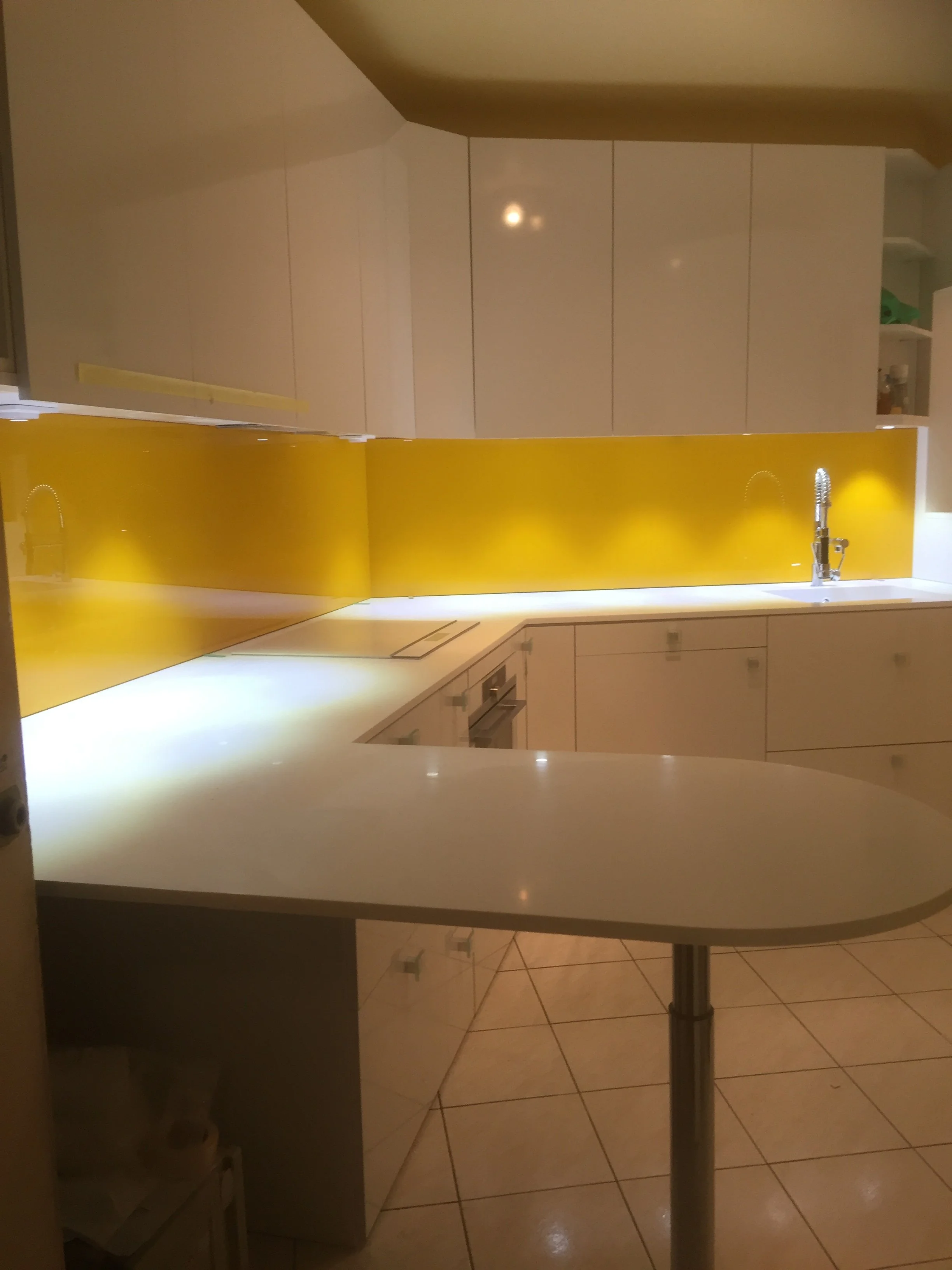 A modern kitchen with a white stacked cabinet, a yellow glass backsplash, and a white countertop. There is a sink with a chrome faucet, and the kitchen floor is tiled with beige square tiles. The overall lighting is warm and cozy.