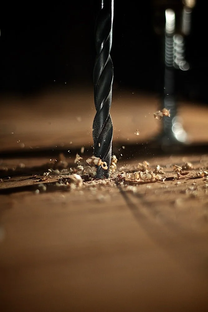 Close-up of a black drill bit drilling into a wooden surface with wood shavings scattered around.