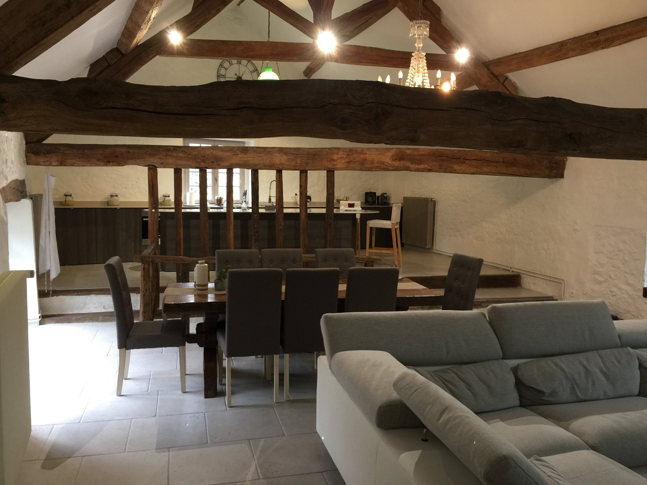 An open-plan interior of a home featuring an upper level with exposed wooden beams and a chandelier. The lower level has a dining table with chairs and a sitting area with a couch. The kitchen is visible in the background with a countertop and window