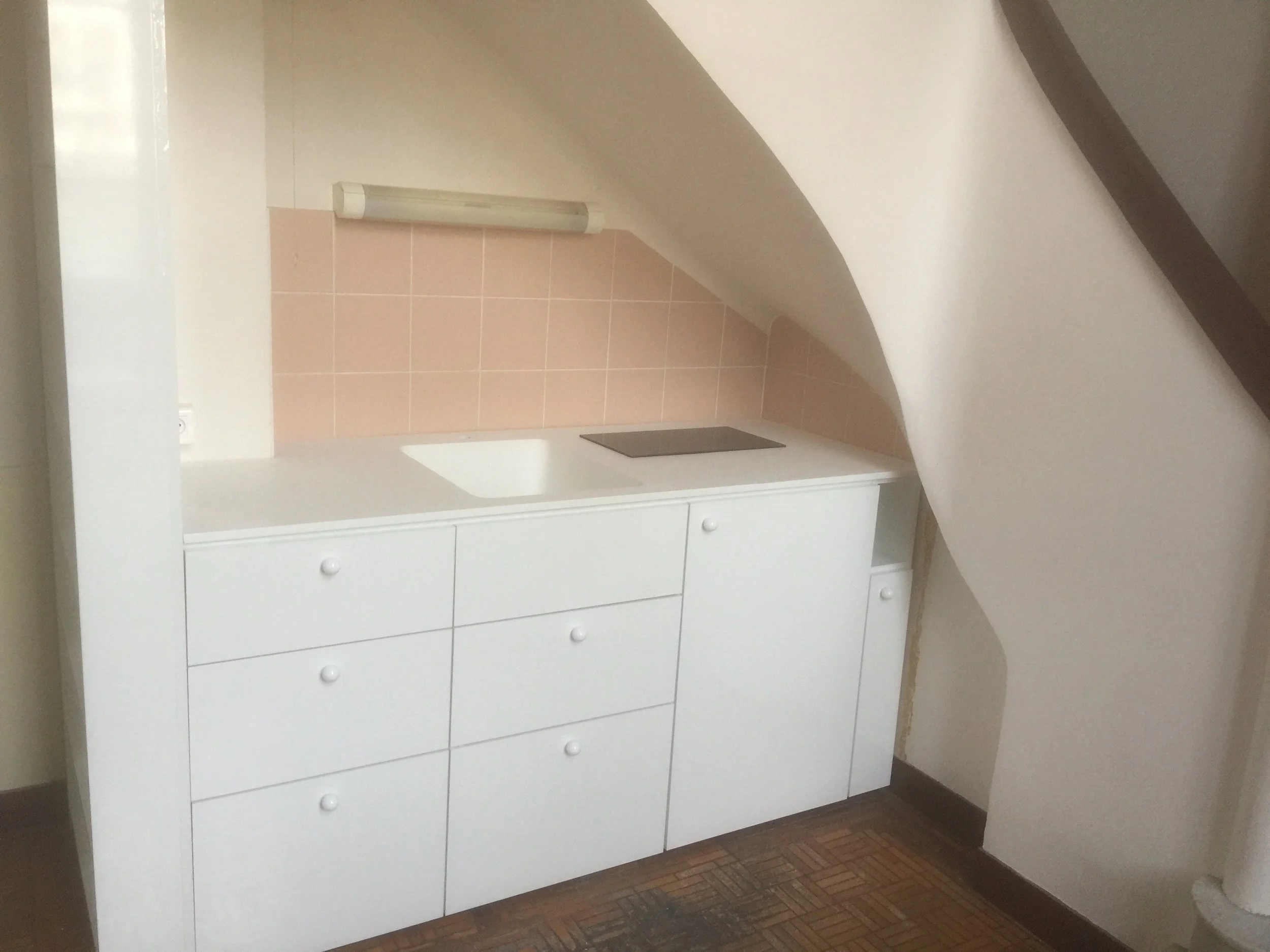 White dresser with multiple drawers and round knobs, situated against a wall with a pink tiled section above and a white countertop. There is a heater or vent on the wall above the dresser and a vent on the floor in front of it.