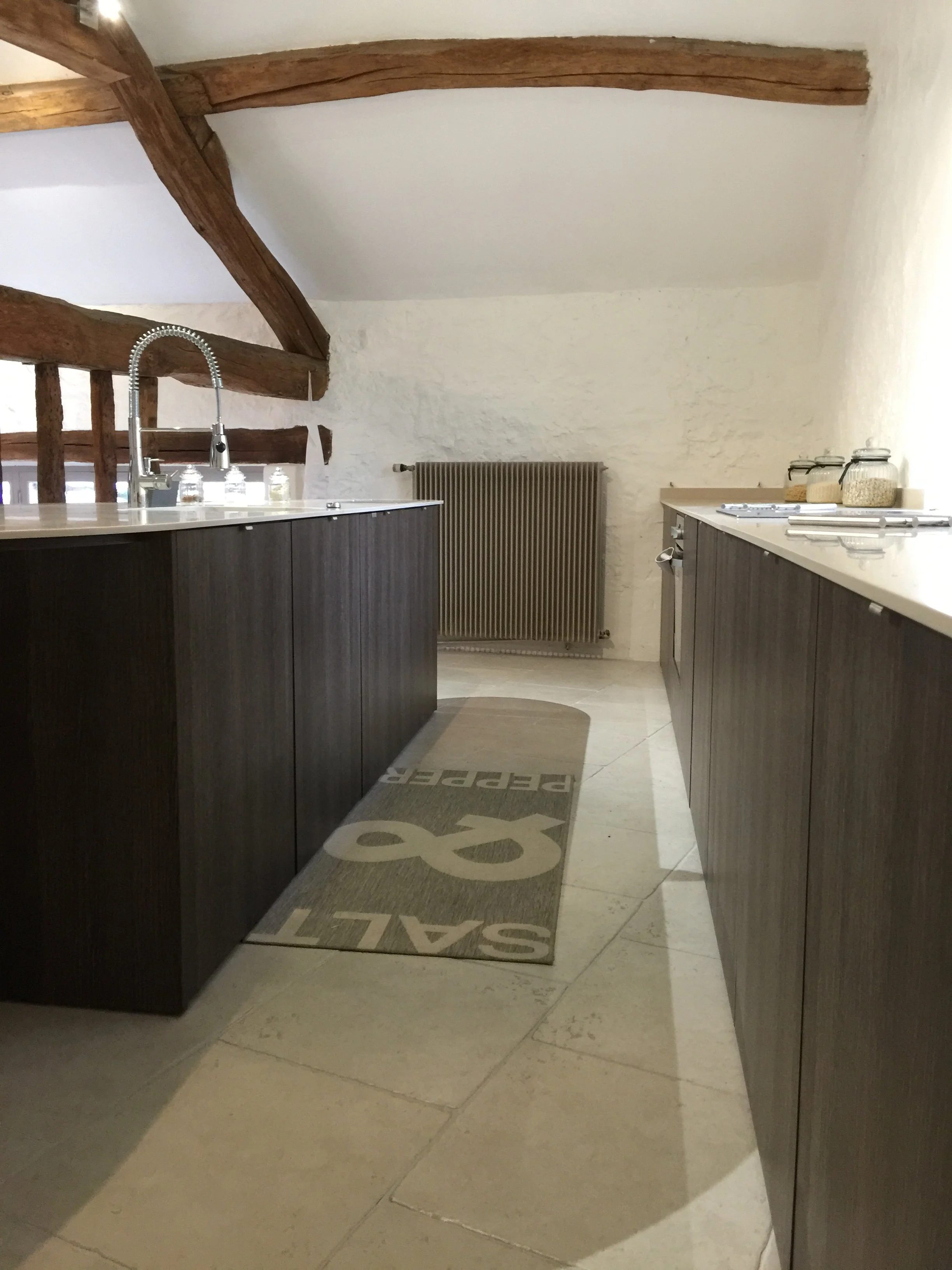 Kitchen area with dark cabinets, a sink with a modern faucet, a radiator on the wall, and jars of grains on the countertop. Wooden ceiling beams and a rug with writing on the tile floor.