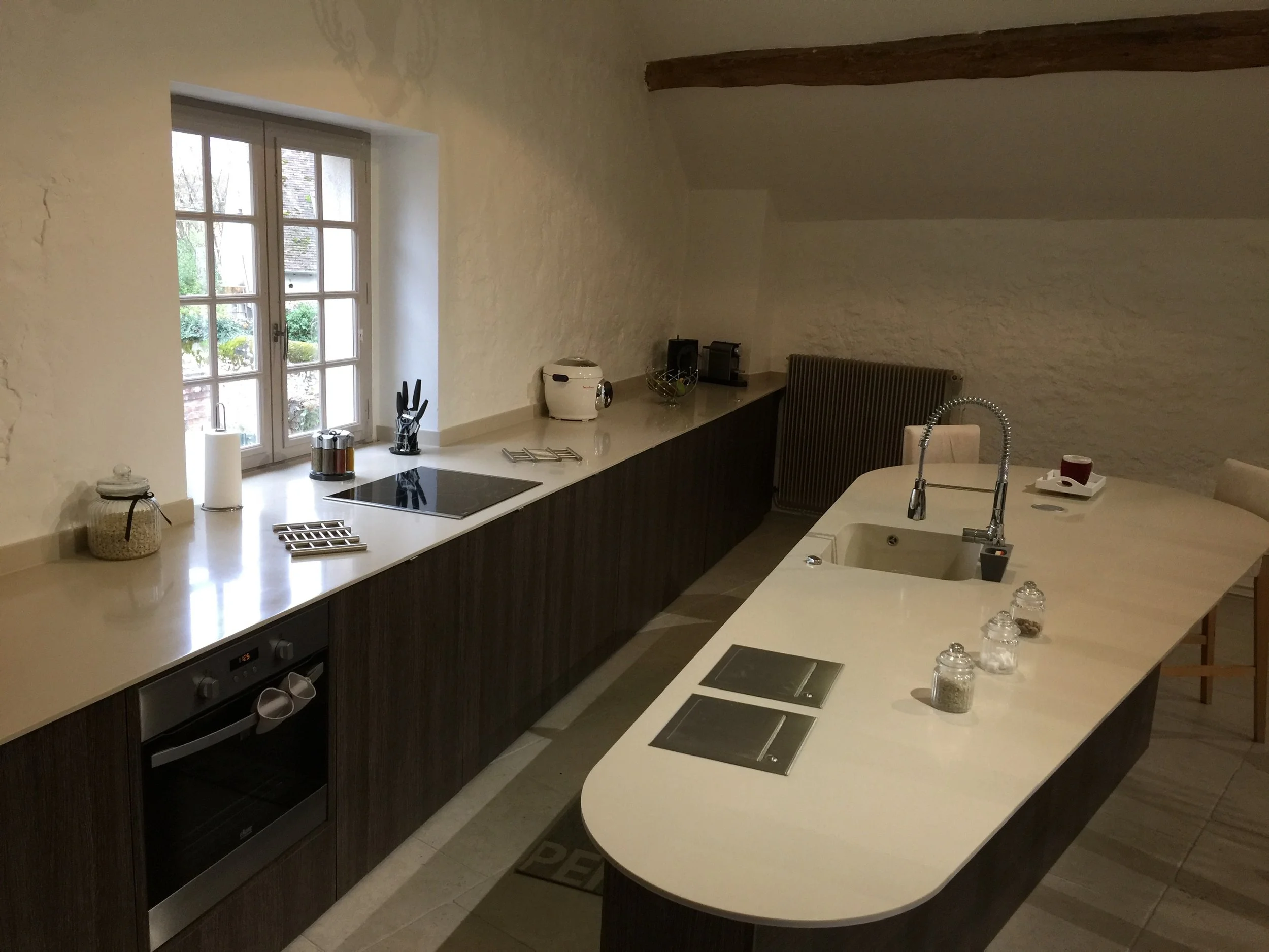 Modern kitchen with white countertops, dark cabinets, a window with a view of outdoors, and various kitchen appliances and utensils.