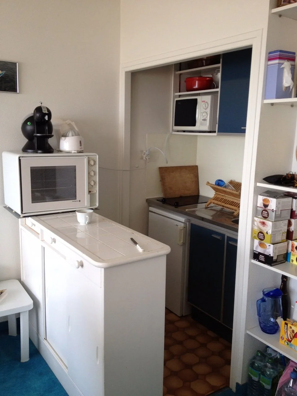 Small kitchen area with microwave, mini fridge, oven, and dish rack, as well as a shelf with food items and a blue pitcher.