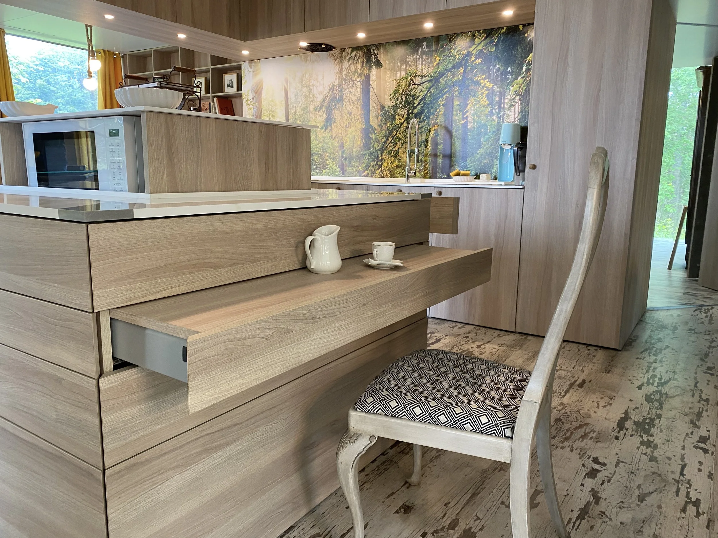Modern kitchen with light wood cabinets and a forest scene mural on the wall, featuring a window with mustard yellow curtains and a chair with a black and white patterned cushion.