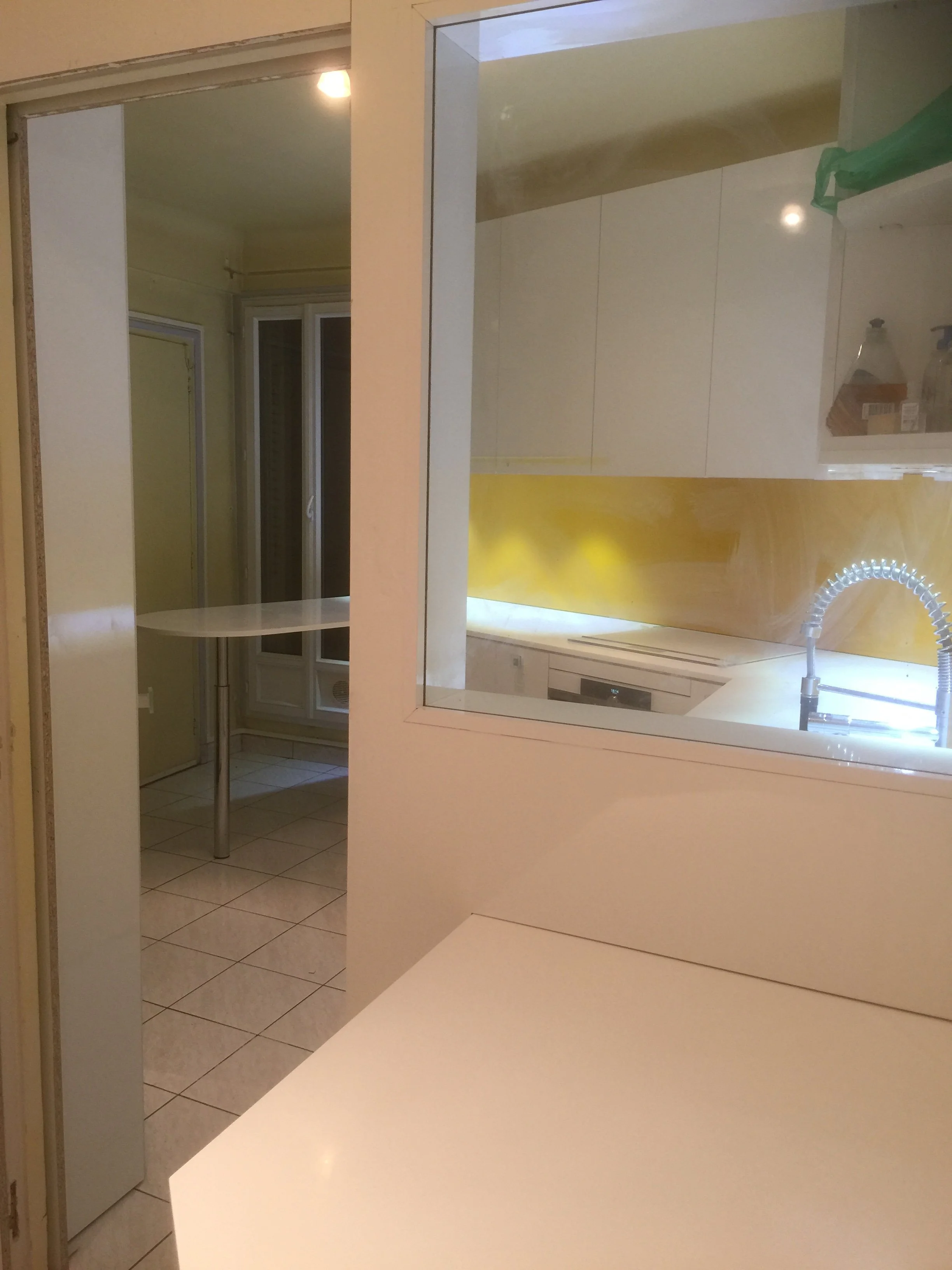 Inside view of a kitchen with white cabinets, a kitchen sink with a flexible hose faucet, and a window. The image is taken from behind a white counter.