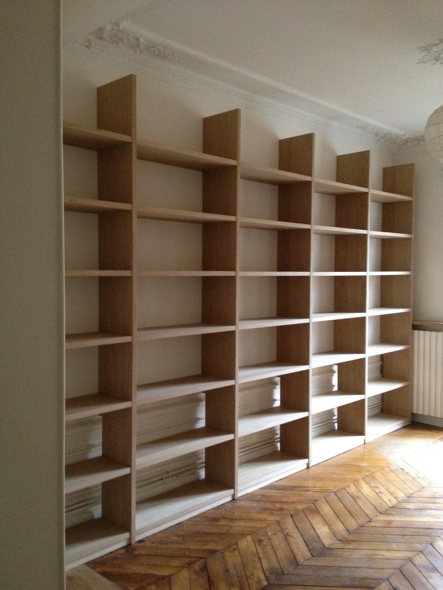 Empty wooden bookshelf with multiple shelves in a room with wooden herringbone parquet floors and detailed ceiling molding.