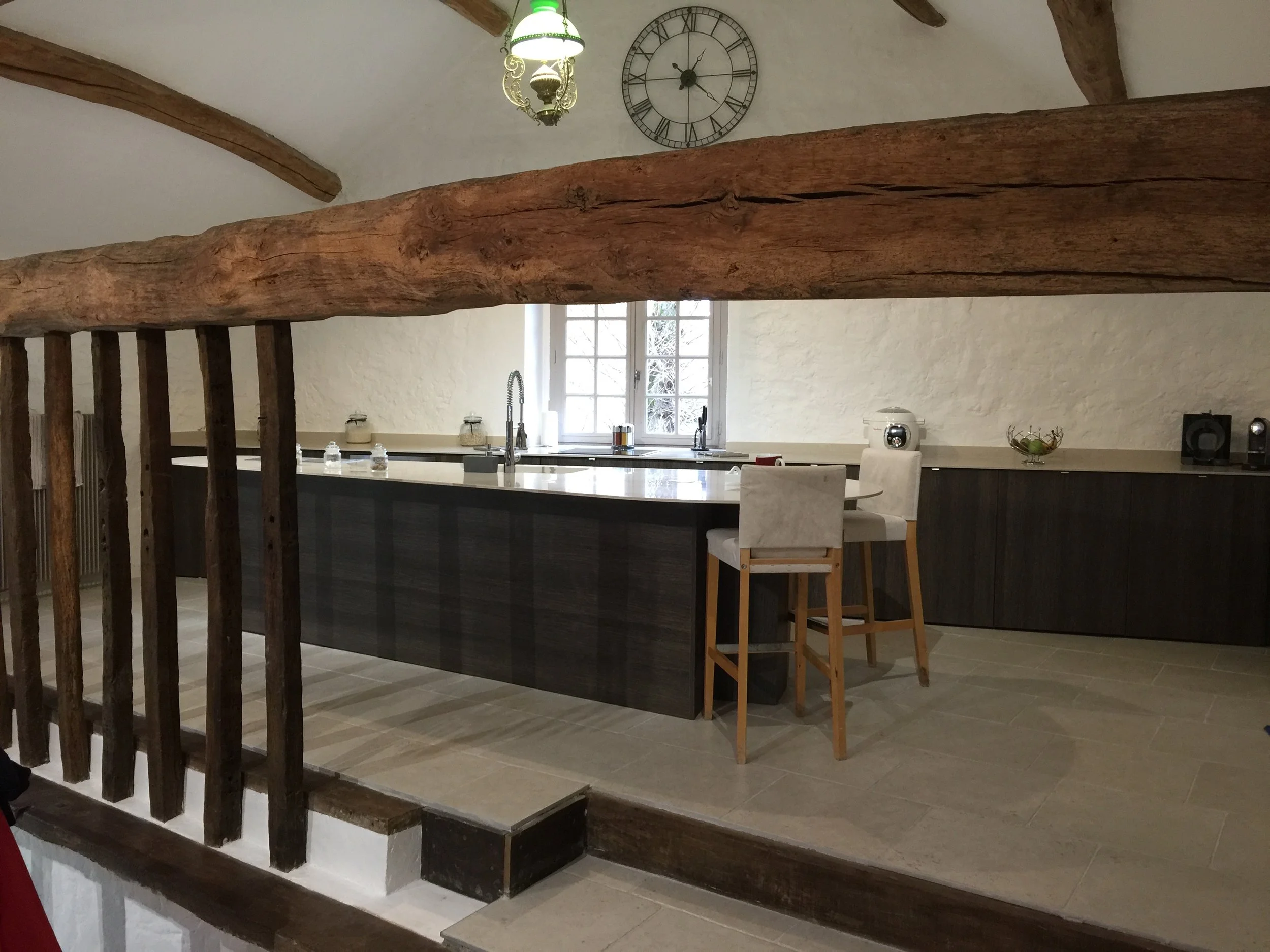 Interior of a house with a rustic wooden beam across the ceiling and a black metal wall clock. There is a kitchen area with a large window, a dark kitchen island, and bar stools. A chandelier with a green light is hanging from the ceiling.