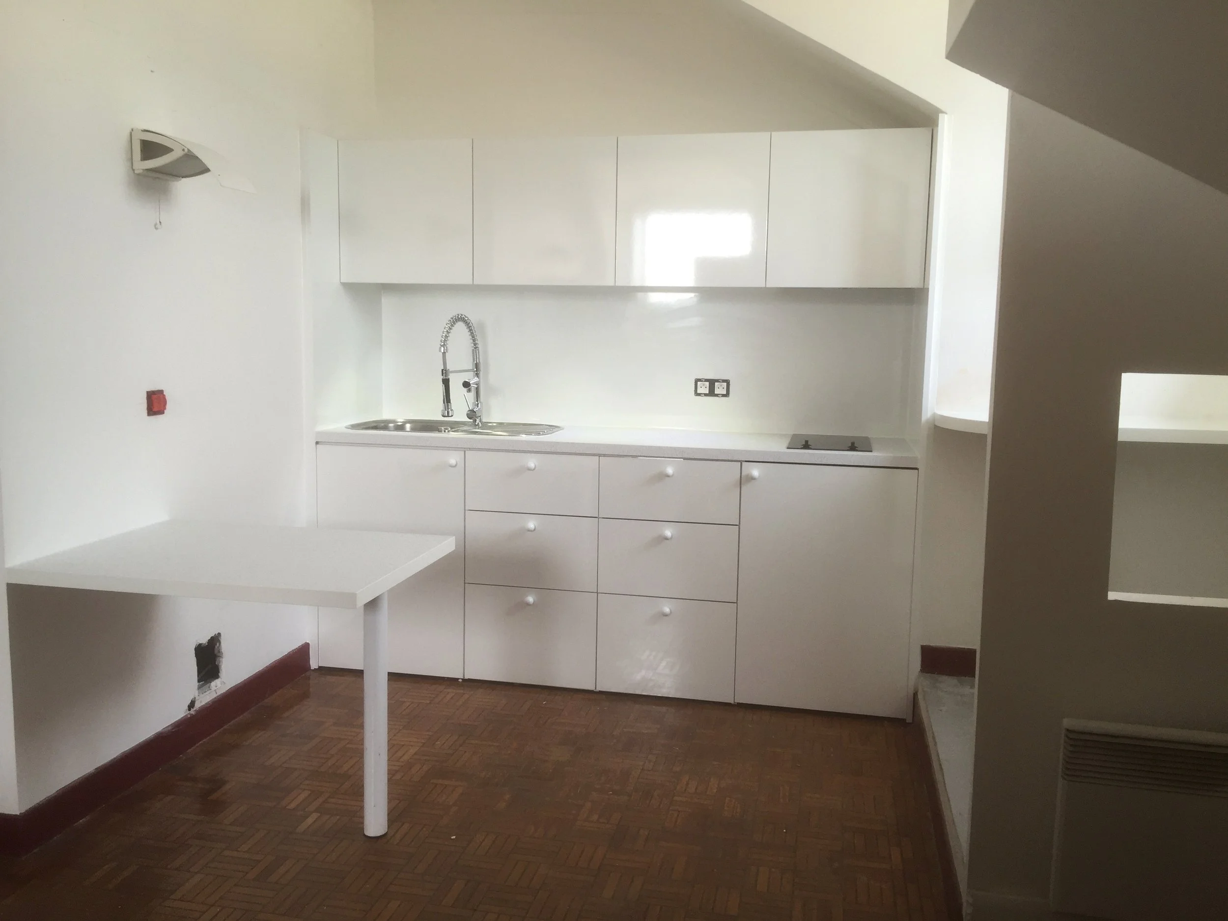 Empty white kitchen with cabinets, a sink with a faucet, and a stove. There is a white table or counter on the left and a hole near the floor on the lower left wall. A power outlet is on the wall above the countertop. The floor is wooden parquet. The