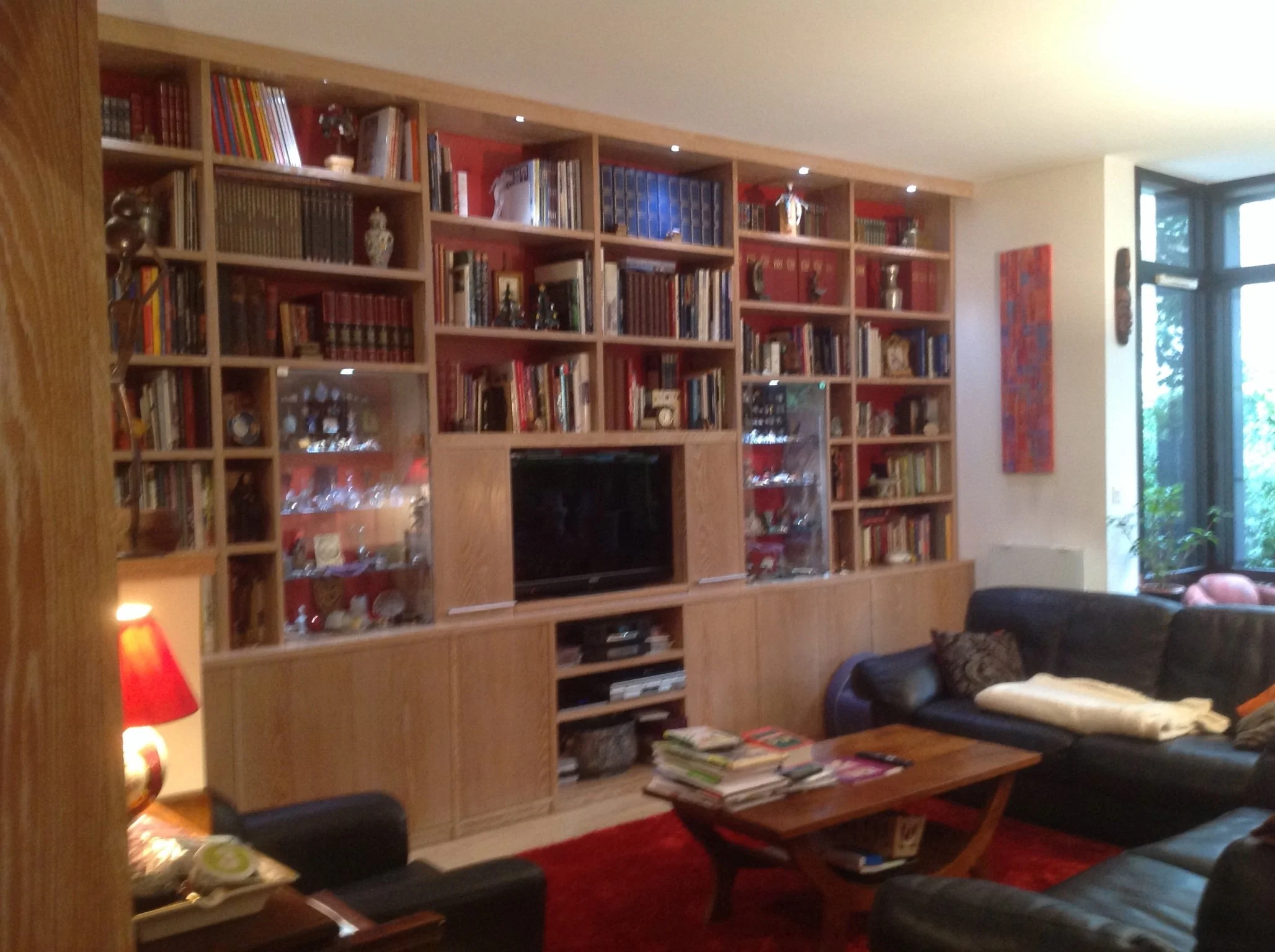 Living room with a large wooden bookshelf filled with books, decorative vases, and ornaments. There is a television in the middle of the shelf, a black armchair with a yellow pillow, and a glass-top table with magazines on a red carpet. A window with